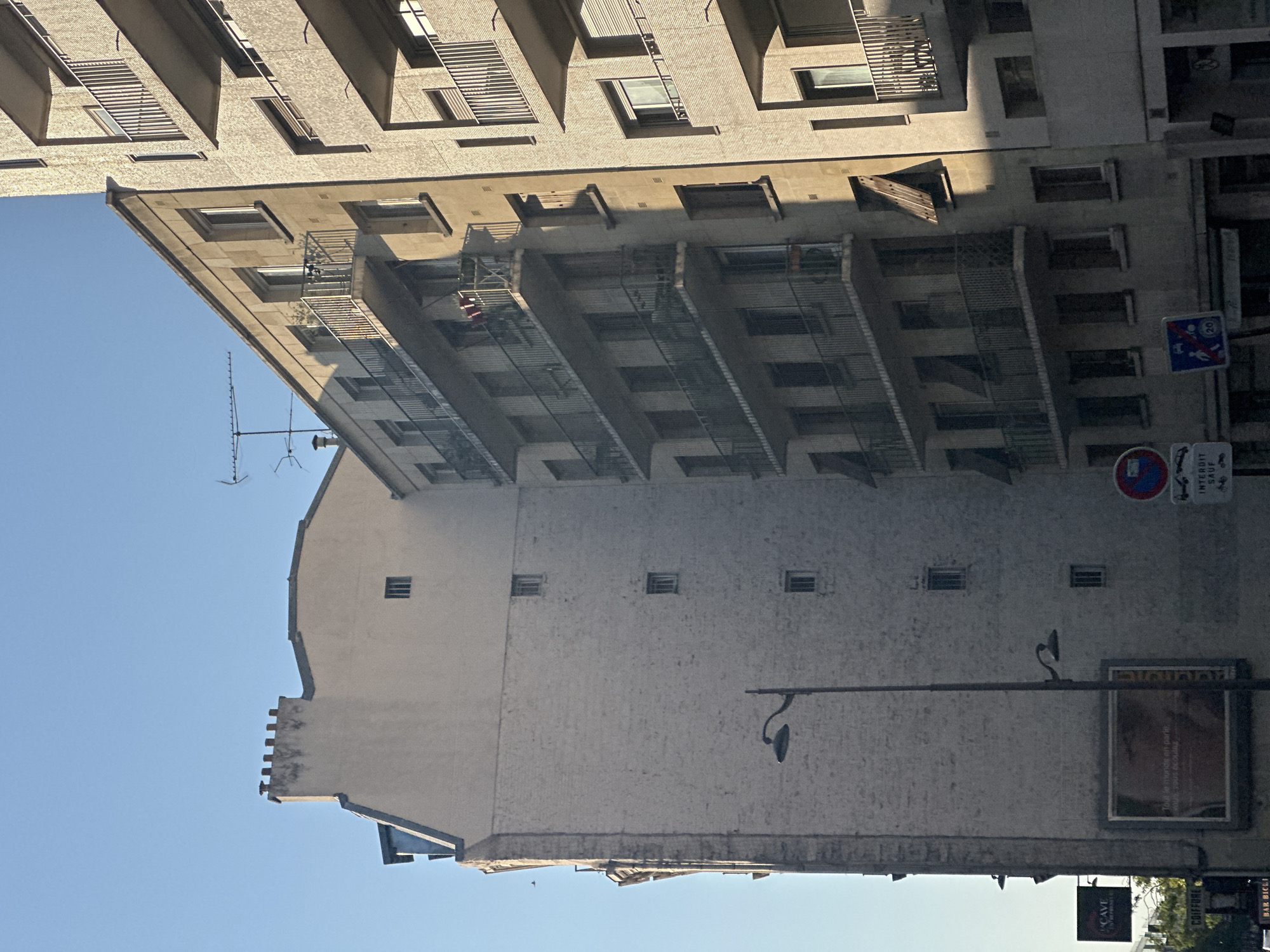 This is a medium-angle, eye-level, full shot of two adjacent apartment buildings on a clear day in Paris, France. The building on the left is older, with a pale beige facade and multiple small, regularly spaced windows. The building on the right is newer and taller, with a textured, pale yellow facade and numerous balconies with railings. The sky is clear and blue. The lower left of the image shows a partial view of a street with trees, a lamppost, signage, and a billboard. The billboard displays a portrait of a person and text. There are no people visible in the image. The architectural style suggests an urban setting in a European city, likely Paris given the presence of characteristic architecture and signage.