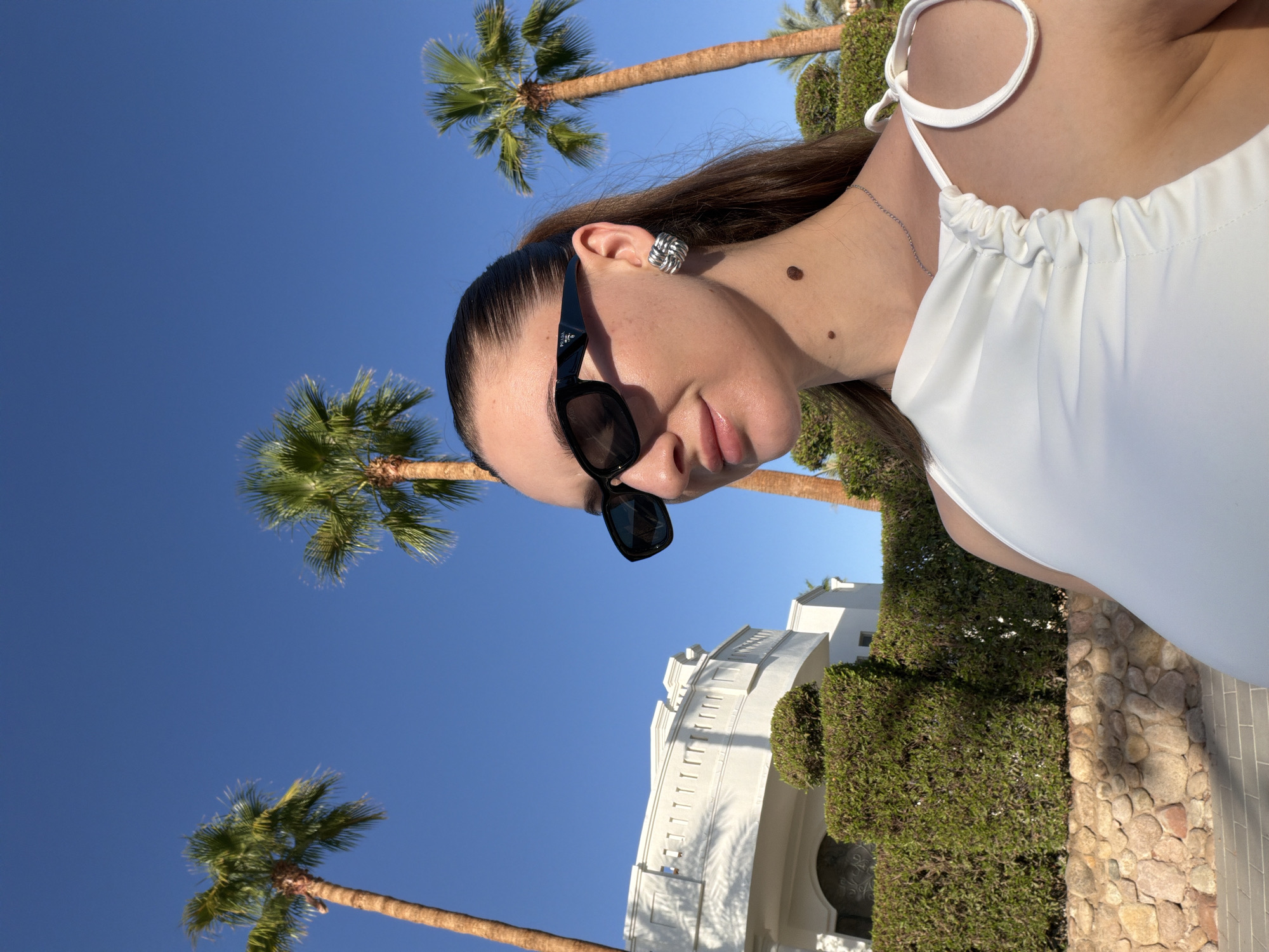 In Sharm El Sheikh, Egypt, a woman with dark hair pulled back and wearing sunglasses and a white top stands in front of a white building with a balcony and manicured hedges. Palm trees are visible against a clear blue sky. The woman has two moles on her neck and is wearing a silver necklace and a large, knot-shaped silver earring. The foreground shows a stone wall.