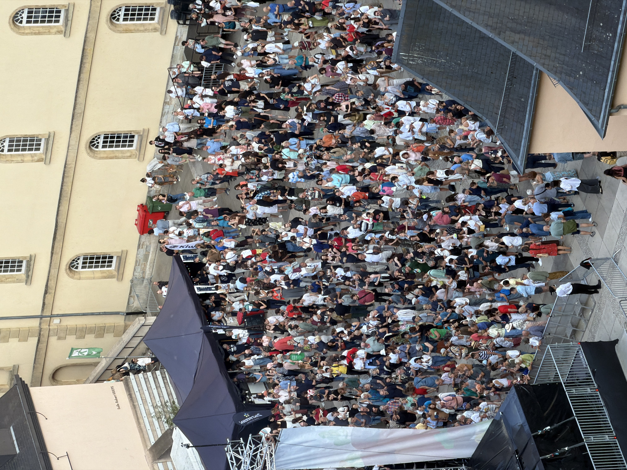 The image captures an outdoor event in Luxembourg City, Luxembourg. The scene unfolds in what appears to be a public square or courtyard, characterized by historic architecture with light-colored facades and arched windows. A large crowd of people, estimated to be in the hundreds, are gathered, suggesting a festival, concert, or public gathering.
In the foreground, a dark tent structure, possibly a stage or vendor stall, is visible, with a white banner hanging from it. A metal barrier with railing separates part of the crowd from the tent. Several individuals are interacting near the tent, some appearing to be staff or performers.
Further back, a group of people are ascending a wide set of stone stairs leading towards a building with the inscription "Salle Robert Krieps" on its facade. A green flag with a white emblem is displayed on a flagpole near the stairs.
The surrounding environment includes other buildings with cream-colored walls and decorative stonework. A red and a green trash receptacle are present near one of the buildings. The ground is paved, indicating a public thoroughfare.
The people in the crowd are diverse in age and attire, with many dressed in casual summer clothing. They are engaged in various activities: standing, walking, talking, and observing. Some individuals are holding phones, likely capturing the event. The overall atmosphere seems to be one of public engagement and enjoyment. The lighting suggests it is daytime, possibly late afternoon or early evening.