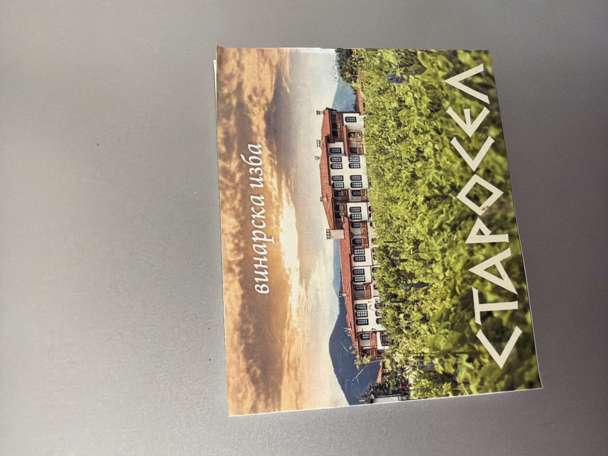 A postcard is displayed on a magnetic surface, showcasing a vineyard and a building under a dramatic sky. The postcard features the text "винарска изба" in white script at the top and "<CTAPOCEЛ" in stylized white block letters at the bottom. The image on the postcard depicts a large, multi-story building with red-tiled roofs and white walls, characterized by numerous windows with dark shutters. This building is surrounded by lush green grapevines, suggesting a winery. In the background, rolling hills rise towards a sky filled with textured, warm-toned clouds. The postcard is positioned slightly askew on a neutral-colored magnetic surface. The location identified is Starosel, a village in the Plovdiv Province of Bulgaria, renowned for its ancient Thracian sanctuary and wineries, and it is not located in the city of Pazardzhik, Bulgaria.