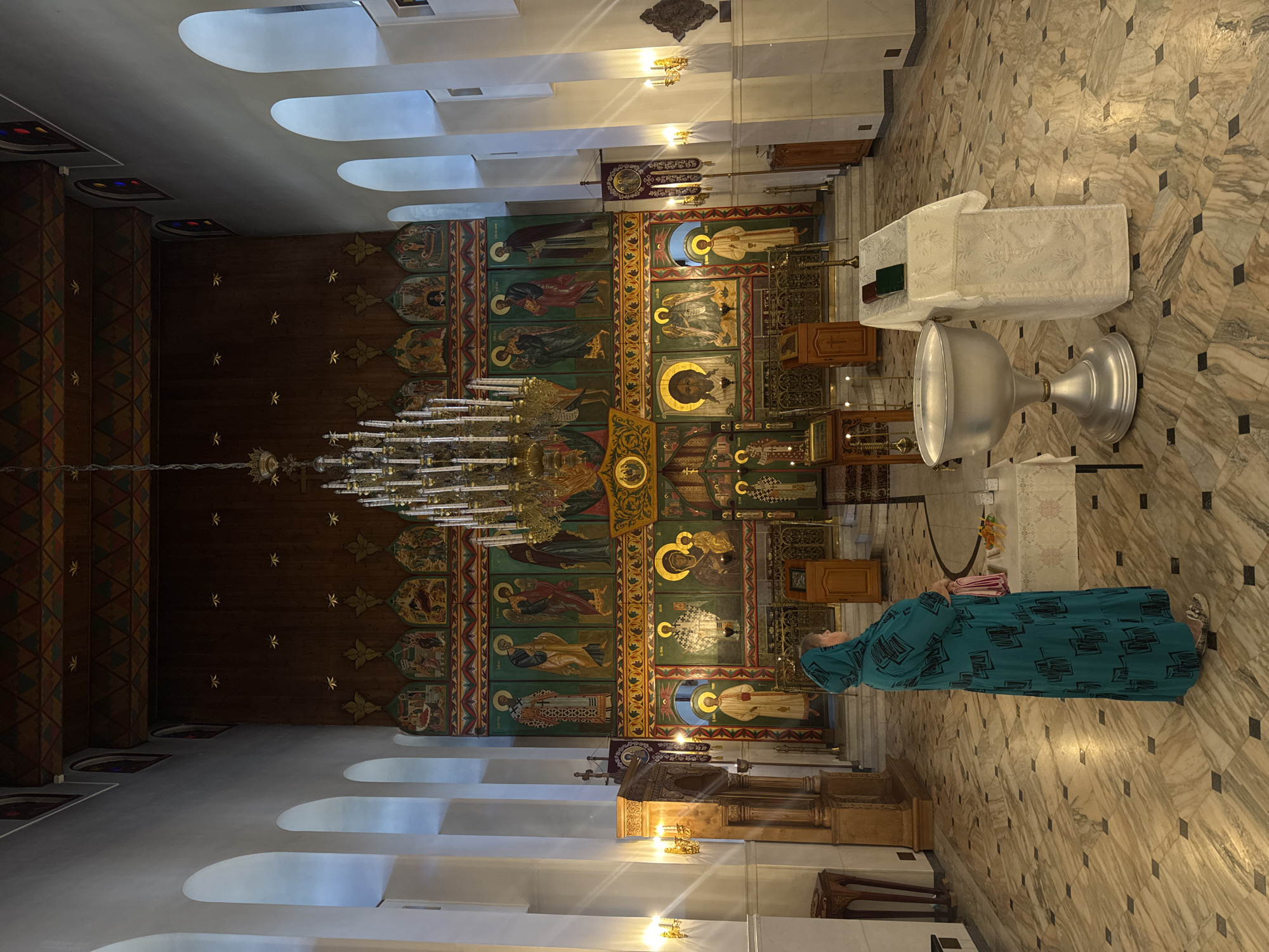 An interior view of the Holy Transfiguration Cathedral in Tolyatti, Russia, captures a woman wearing a teal headscarf and a matching long dress with a black geometric pattern, standing in prayer. She is positioned in the foreground, facing away from the camera, her right hand resting on a small table draped with a white cloth. The cathedral's interior is richly adorned with religious iconography. Dominating the rear wall is an iconostasis, a richly decorated screen featuring multiple tiers of icons depicting religious figures. A large, ornate chandelier hangs from the ceiling, illuminating the space. The floor is tiled with a marble-like pattern of light and dark squares and rectangles. In the mid-ground, several liturgical objects are visible, including a large silver baptismal font, a wooden pulpit, and a table with candles. The walls are white and feature arched windows, and the ceiling is adorned with wooden panels and decorative elements. The overall atmosphere is one of solemnity and religious devotion.