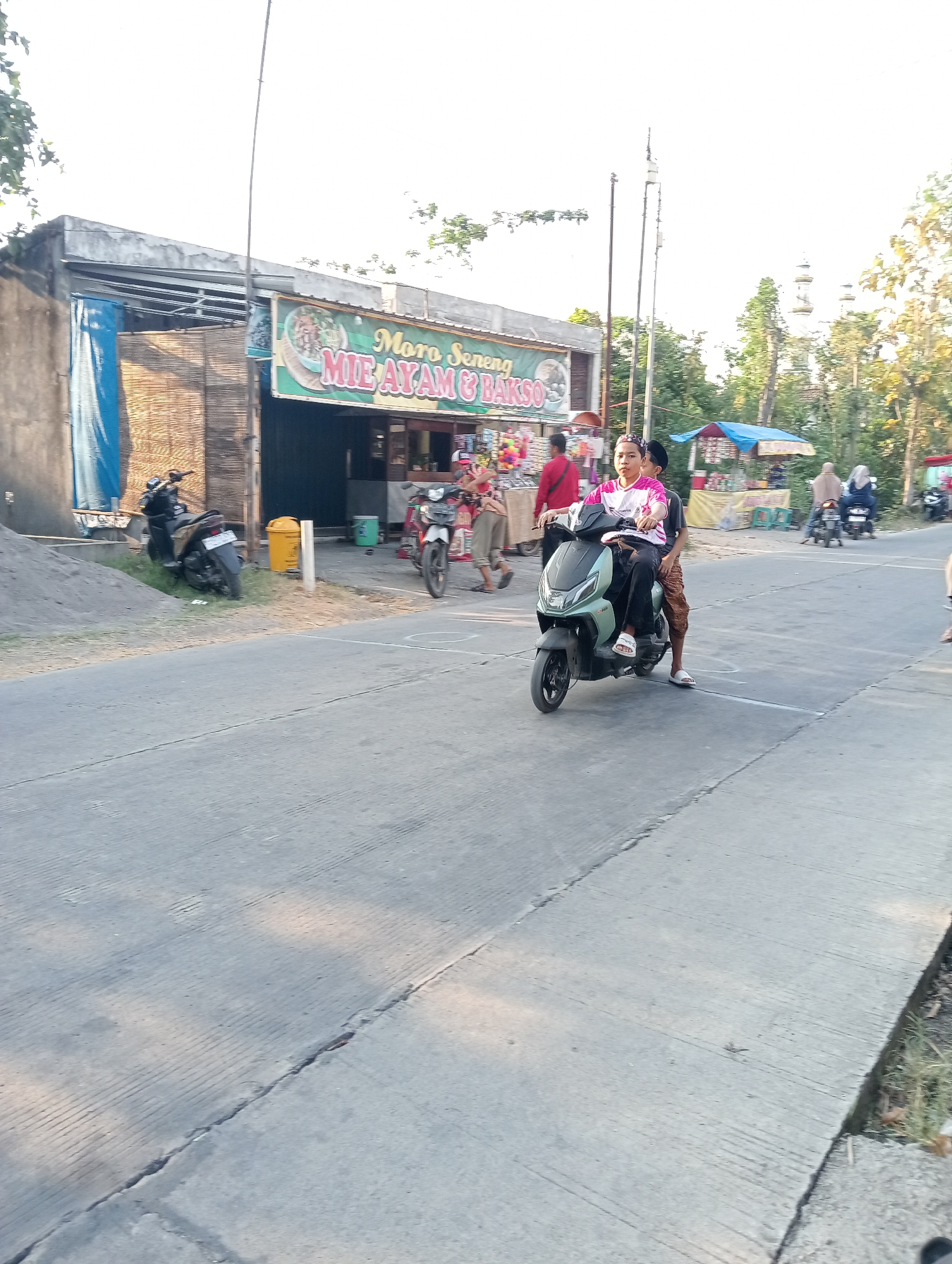 The image depicts a street scene in Tanjunganyar, Indonesia, with a focus on a food stall and passing vehicles. In the foreground, a paved road with distinct parallel grooves stretches across the frame. A light green scooter carrying two young individuals, both males, is moving from left to right. The rider, wearing a white and pink shirt, is in control, while the passenger, in a dark shirt, sits behind him. Both are wearing casual clothing.
To the left, a food stall with a green awning displaying "MIE AYAM & BAKSO" (Chicken Noodles & Meatballs) is visible. Behind the stall, a section of a building with a textured wall and a blue tarp is present. A black motorcycle is parked on the grassy area beside the stall. A yellow trash bin stands near the entrance of the stall.
Further down the road, other vehicles and people are scattered. A white motorcycle with a rider is seen in the middle ground to the right of the food stall. In the background, trees and some structures, possibly including minarets, are visible, suggesting a mix of urban and semi-rural surroundings. The lighting indicates it is daytime, likely late afternoon, with soft shadows cast across the scene.