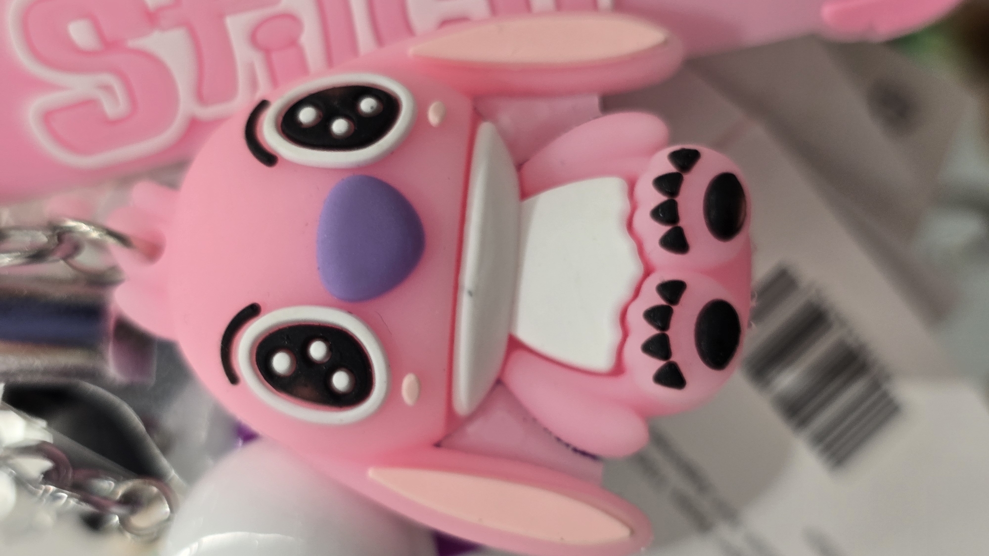 This is a close-up, eye-level, vertical shot of a pink, rubber keychain in the shape of a cartoon character, possibly Angel from Lilo & Stitch. The character has large, black, oval eyes with white highlights, a purple nose, and pink ears. Its body is pink with a white chest area and black paw prints on its feet. The background is blurred and out of focus, showing hints of other objects, possibly in a retail setting, such as a barcode and a pink item with embossed white lettering. There is no discernible location in Krasnodar, Russia, or any people present.