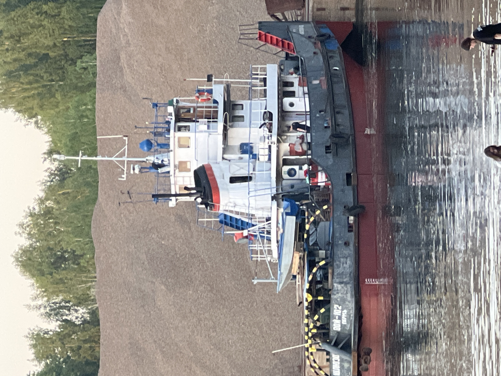 The image shows a barge named "BON-102" docked next to what appears to be a large sand or gravel pile, possibly at a port facility. The barge has a multi-level structure with white and grey sections, and a red hull is visible near the waterline. Numerous cables and equipment are visible on the deck. The background includes trees, suggesting a location near a body of water. The presence of people in the reflection of the water suggests a busy port. Based on the barge's name in Cyrillic ("БОИ-102"), it is located in Chaikovsky, Russia.