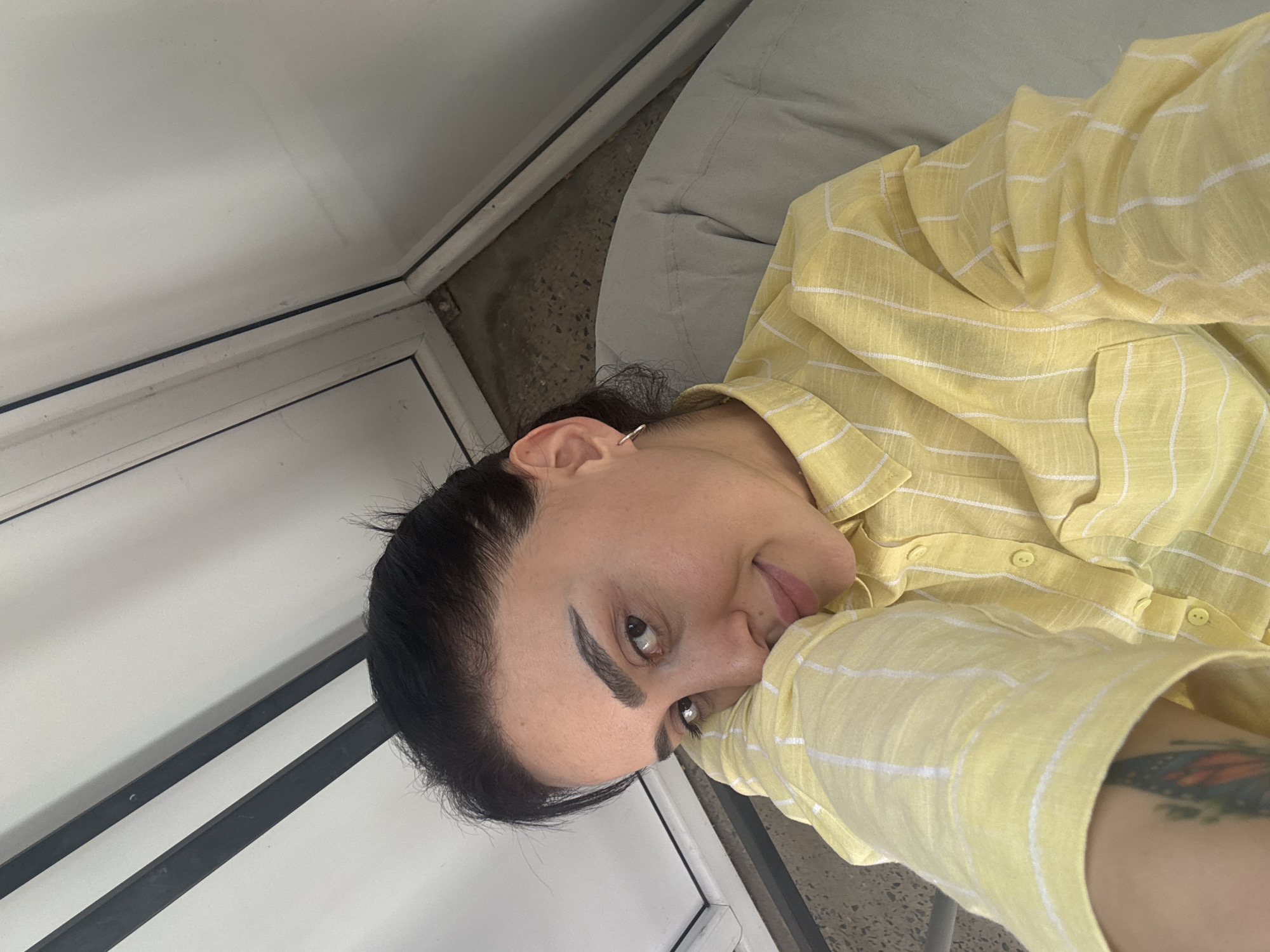 A person is seen in a close-up, angled shot. They are wearing a yellow and white plaid button-down shirt. Their dark hair is pulled back, and they have distinct, thick eyebrows. The person is smiling slightly and their gaze is directed towards the camera. A small gold hoop earring is visible on their left ear. A tattoo can be seen on their right arm. The background includes a light gray cushion and a white door or wall with a black handle or frame. The location of Tbilisi, Georgia is not discernible from the image.