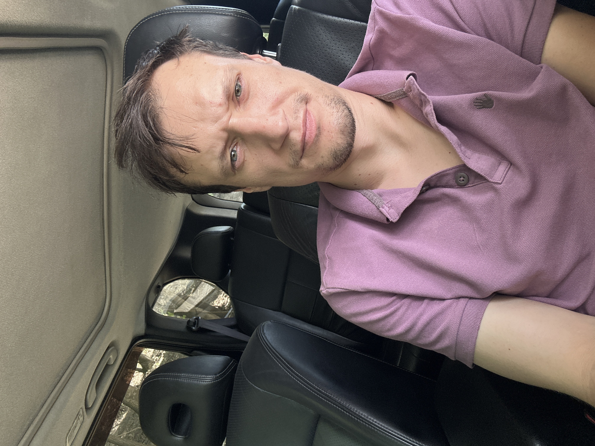 A man with light skin and brown hair is sitting in the front passenger seat of a car. He is looking at the camera with a stern expression. He is wearing a purple polo shirt. The car's interior is visible, including black leather seats and a sunroof. Outside the car, trees and foliage can be seen through the window, suggesting an outdoor setting, possibly in a rural or wooded area. No specific location in Makiv, Ukraine, can be determined from the image.