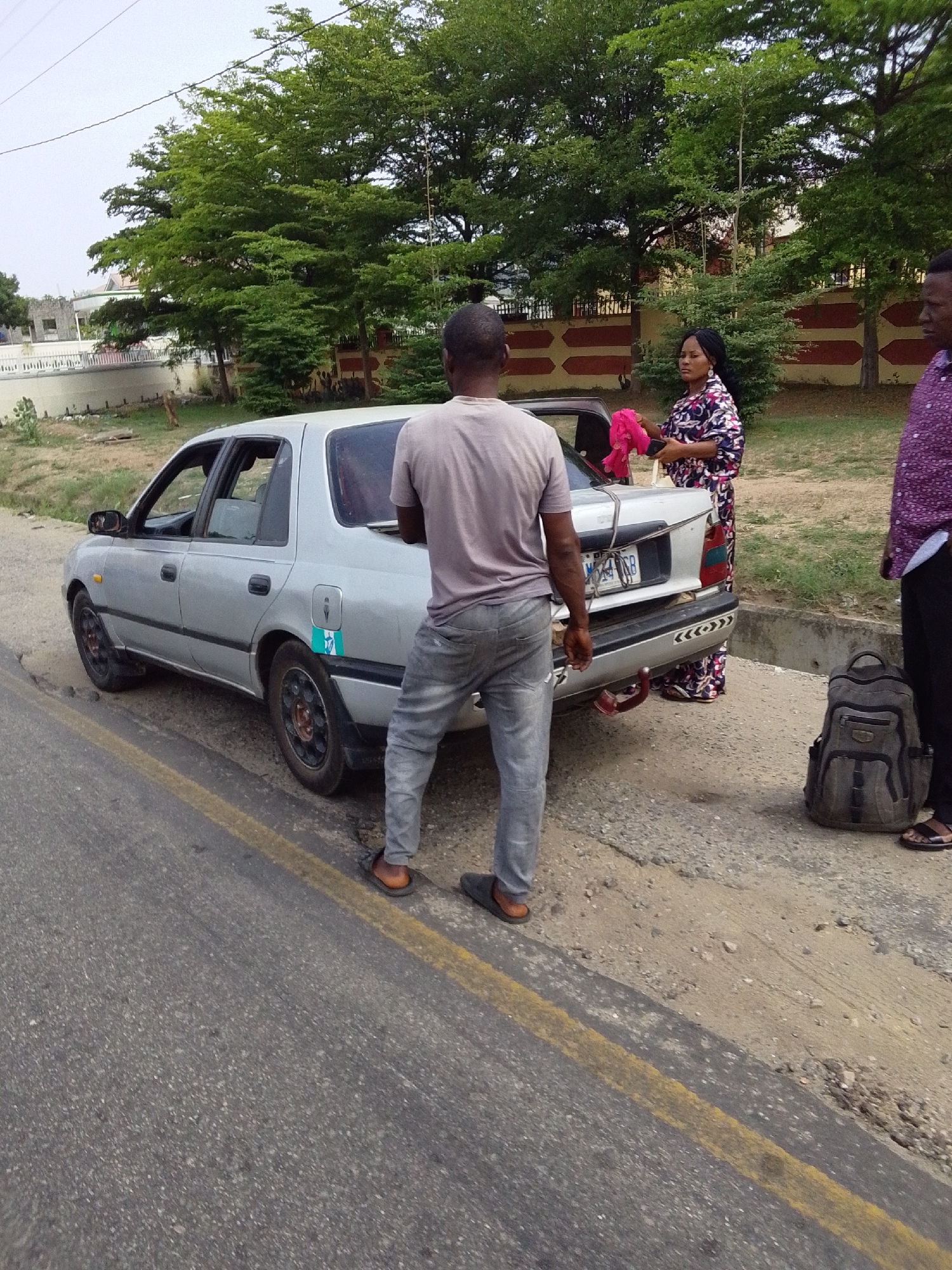 A silver sedan with a dent in the rear passenger-side door sits on a roadside in Lokoja, Nigeria. A man in a t-shirt and jeans stands near the car's trunk facing away from the camera. A woman in a patterned dress holds a pink cloth behind the car. A third man in a purple patterned shirt and black pants stands beside a gray backpack. Trees and a wall are visible in the background. The road has a yellow line marker.