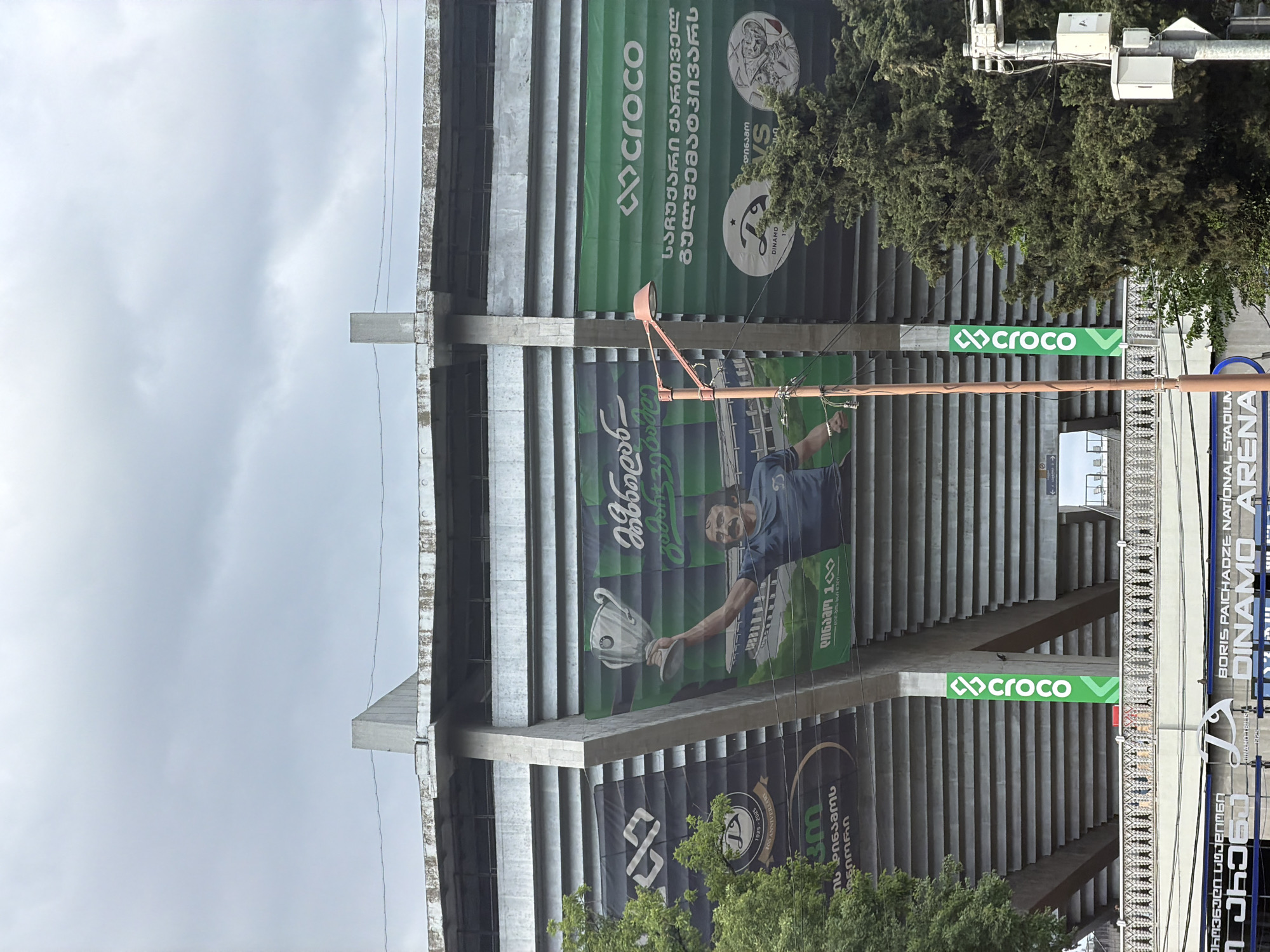 The image shows a section of the Boris Paichadze National Stadium in Tbilisi, Georgia, also known as the Dinamo Arena. The stadium's concrete structure is visible, with various banners and advertisements attached to it. One banner displays a soccer player holding a trophy. The "croco" logo is prominently displayed on several green signs. A street lamp is also in view, attached to the stadium's structure. Trees are visible around the stadium, and the sky is overcast. Banners display slogans in Georgian.