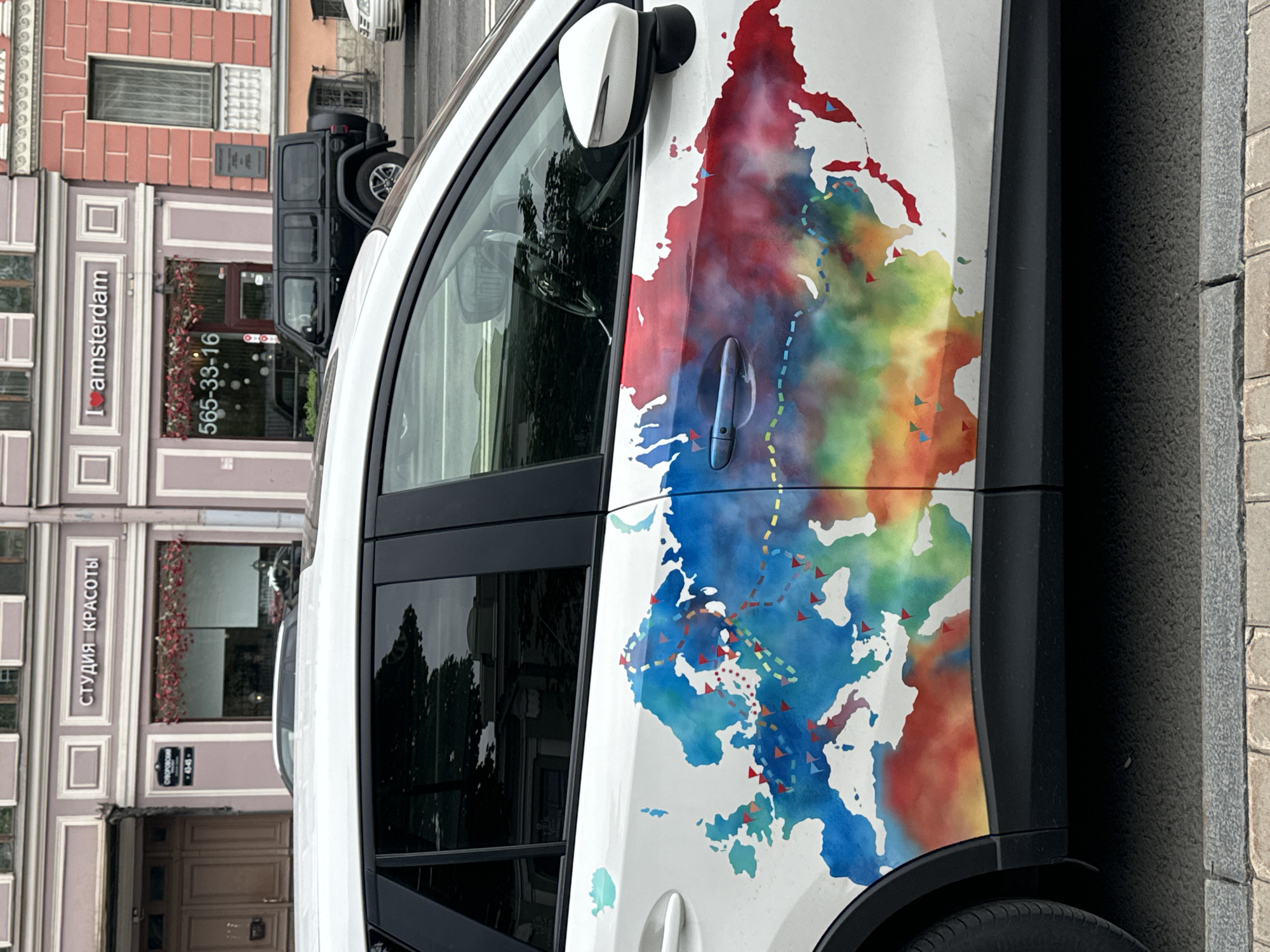 A white SUV is parked on a city street in Saint Petersburg, Russia. The side of the SUV is decorated with a watercolor map of Eurasia, featuring colorful gradients and small red and blue triangular flags marking various locations. The street is paved with cobblestones and bordered by a sidewalk. In the background, a building with pink and purple facades features several storefronts with signage in Russian, including one that reads "СТУДИЯ КРАСОТЫ" (Beauty Studio) and another that says "I amsterdam". A black Jeep Wrangler is parked in front of one of the storefronts.