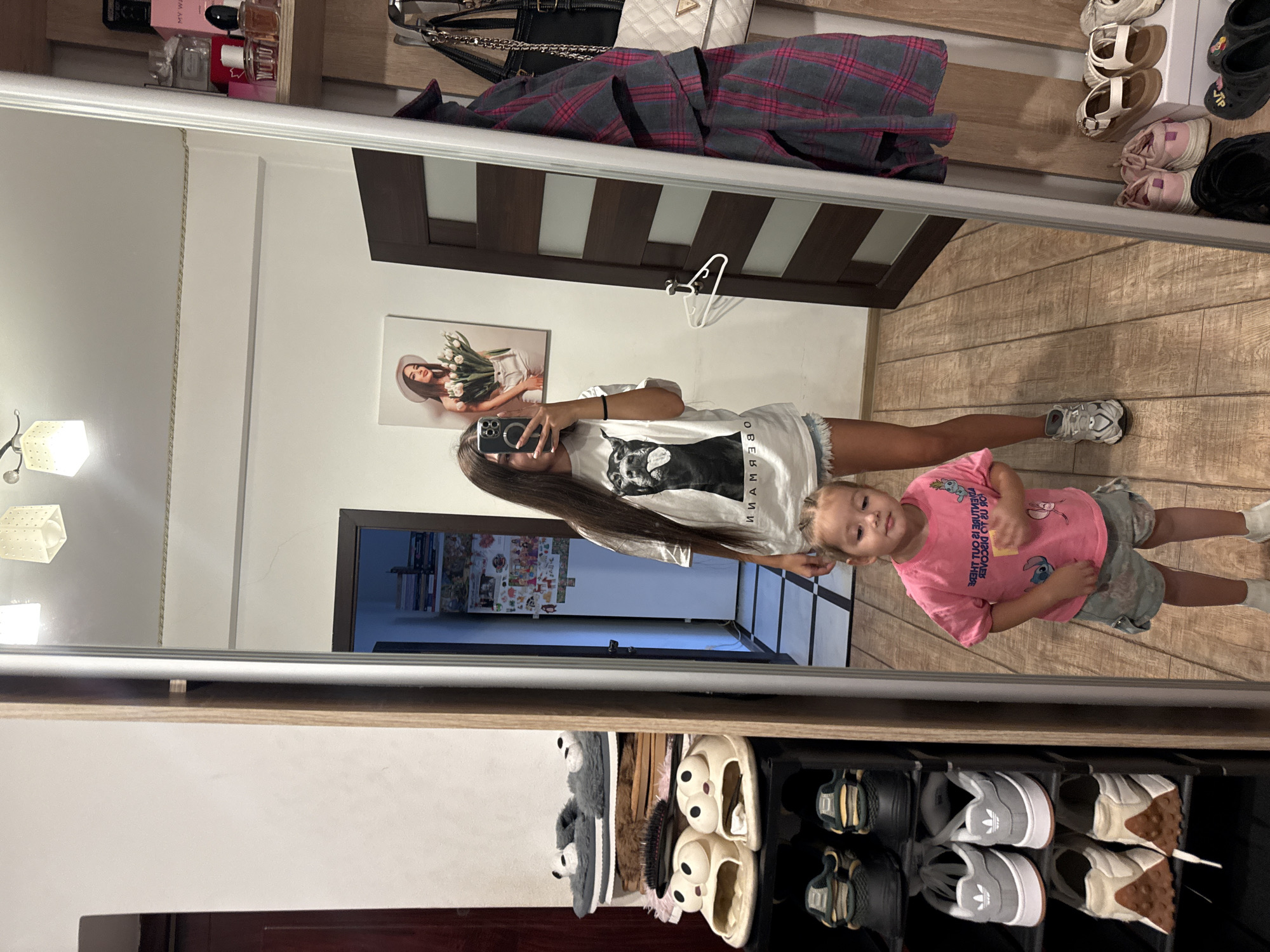 This is a full-length, medium shot photograph taken inside a residential room, likely a hallway or dressing area, in Chișinău, Moldova. The room features a large mirror that reflects most of the scene.
In the foreground, a young girl, appearing to be around 3-4 years old, stands in the center of the frame, facing the camera. She has braided hair and is wearing a pink t-shirt with cartoon characters and grey camouflage-patterned shorts. She is looking down with a slightly tilted head.
To the left of the frame, a shoe rack is visible, filled with various footwear. On the top shelf, there are two pairs of fluffy grey slippers with panda-like faces. Below that are other slippers and on the lower shelves, several pairs of athletic shoes, including a pair of grey Adidas sneakers.
Reflected in the mirror on the left side is a young woman, likely the older sister of the child. She has long, dark hair and is wearing a white t-shirt with a graphic of a dog and denim shorts. She is holding a smartphone and taking a selfie.
To the right of the woman in the mirror, a dark brown wooden door is visible, with a white hanger attached to it. Further to the right, a section of a wardrobe or shelving unit is present, displaying folded clothing, handbags, and perfume bottles. A framed picture of a woman holding tulips hangs on the wall above the door.
The floor is made of light brown wooden planks laid in a parquet pattern. The lighting in the room appears to be from overhead fixtures, one of which is visible in the top center of the mirror reflection. The overall impression is of a personal, lived-in space.