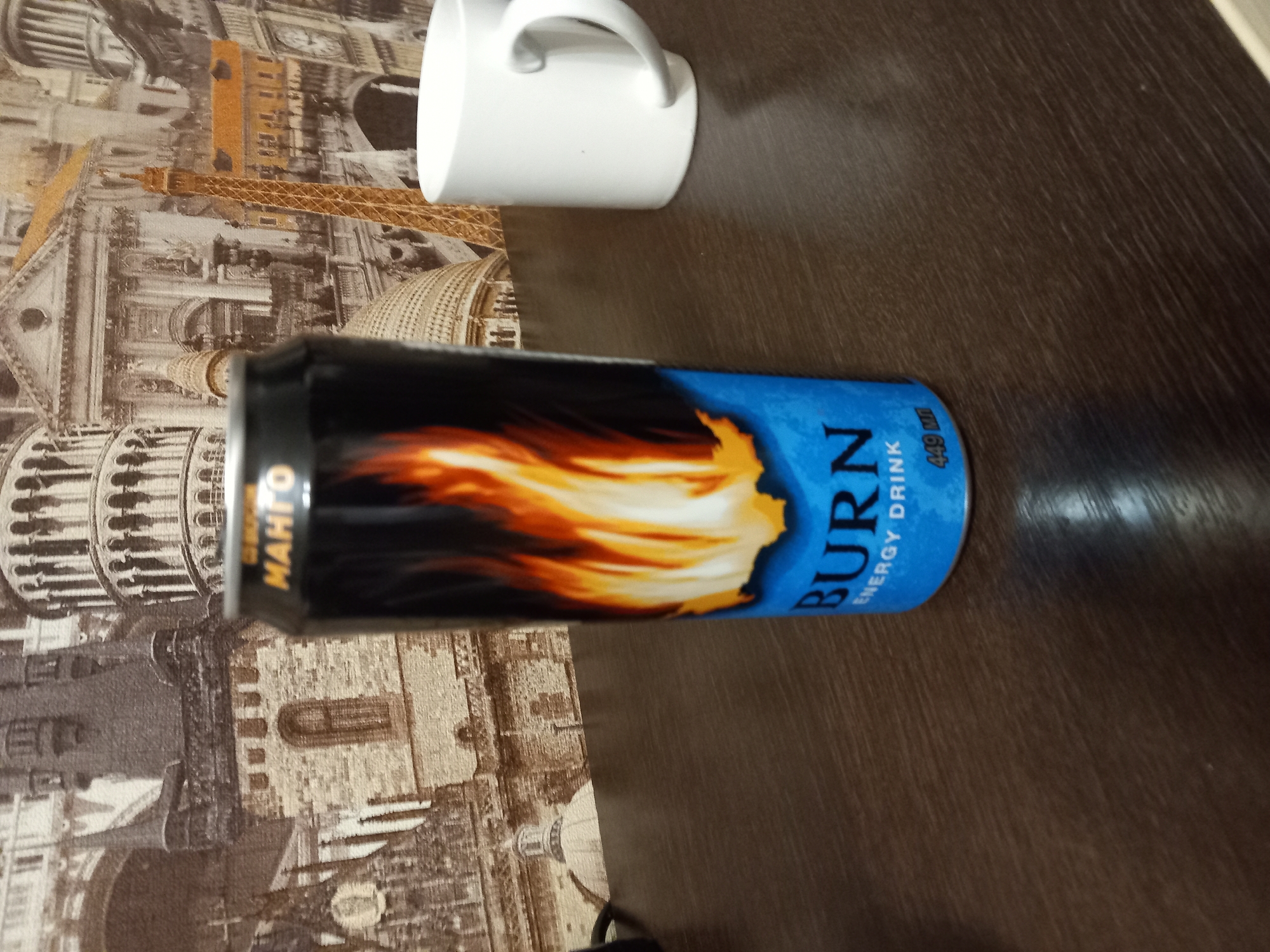 The image shows a close-up, eye-level shot of a "Burn" energy drink can placed on a dark wooden desk. The can is predominantly black with a design of orange and yellow flames. The word "BURN" is prominently displayed in large, white, sans-serif font on a bright blue background at the bottom of the can, followed by "ENERGY DRINK" and "449 ml". To the right of the can is a white ceramic mug. The background is a wall covered in a wallpaper featuring a collage of European landmarks in sepia tones, including what appears to be the Eiffel Tower, the Leaning Tower of Pisa, and architectural elements reminiscent of Rome. The lighting is dim, and the focus is sharpest on the energy drink can.