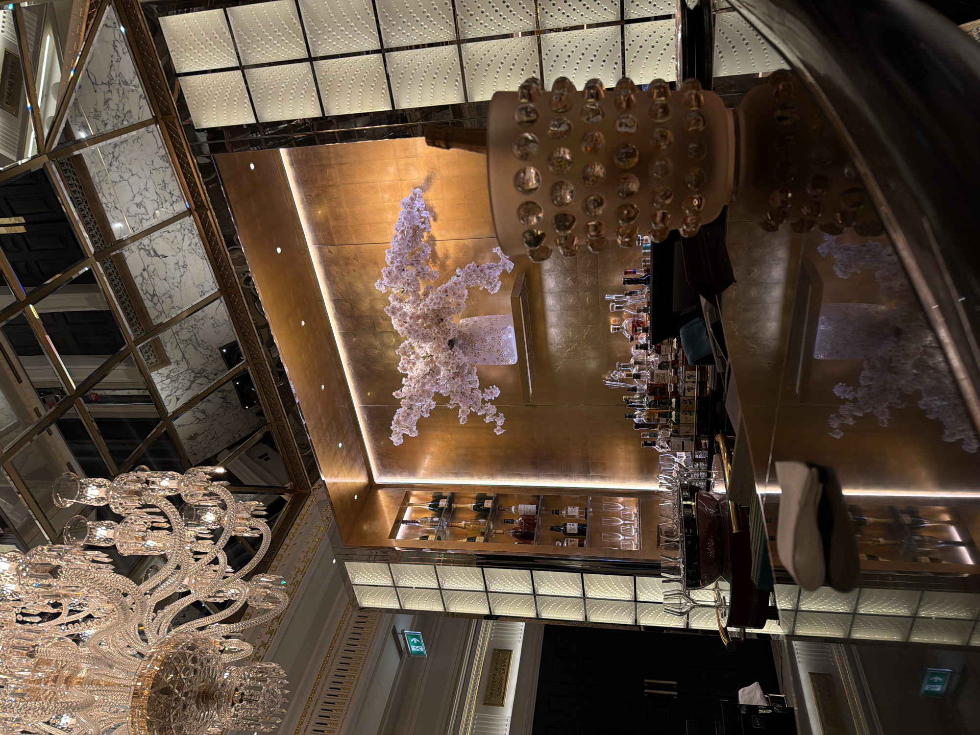 The image is an interior shot of a high-end establishment in London, United Kingdom. A large, ornate crystal chandelier is a focal point, complemented by a smaller floral arrangement. The decor features gold accents and mirrored surfaces, adding to the luxurious ambiance. Shelves stocked with various bottles of alcohol suggest a bar or lounge setting. The lighting is soft and warm, and a patterned ceiling is visible. An exit sign is present, and glasses and other bar equipment are visible on a countertop.