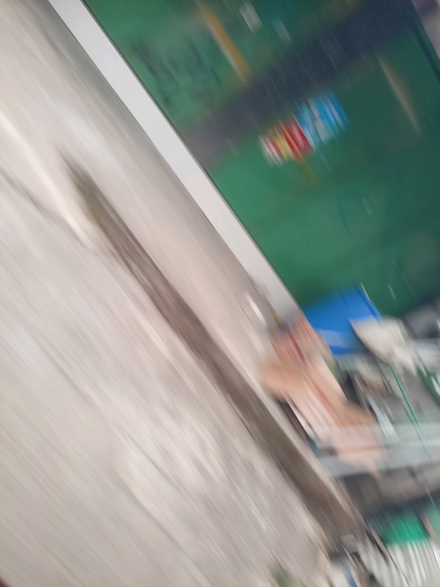The image is a blurry, diagonal shot of a space that appears to be an indoor area with objects and surfaces. The upper portion of the image features a green surface with faint, multicolored shapes that could be signage or graphics. A white line separates this green area from a lighter, textured surface that occupies the majority of the frame. This textured surface appears to have a pattern of parallel lines or grain, suggesting it might be a wall or a large object. In the lower right quadrant, there are hints of other objects, including a bright blue rectangular shape and some lighter colored items that are difficult to discern due to the blur. The overall impression is one of motion blur, making specific identification of objects and their functions challenging. The provided context of "Kebonlegi, Indonesia" does not offer any specific visual cues within the image itself to confirm or deny this location.