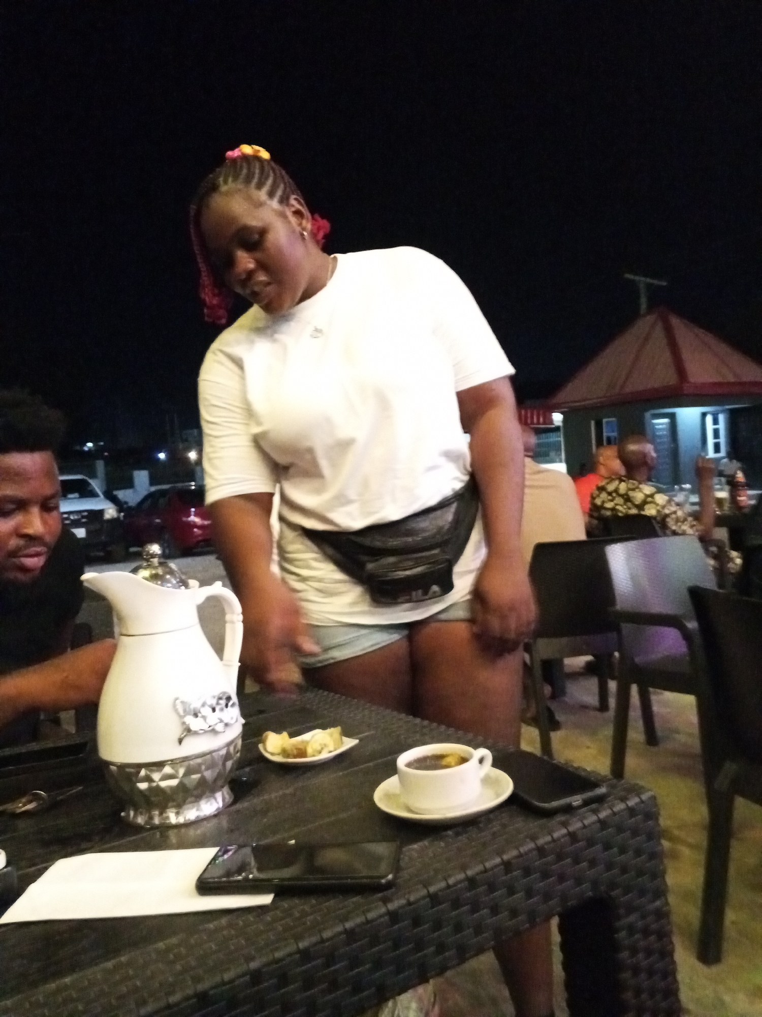 The image depicts an outdoor scene in what appears to be Lokoja, Nigeria, at night. A Black man and a Black woman are present. The woman, in a white T-shirt, denim shorts, and a Fila waist bag, stands near a table. The man, in a dark shirt, is seated at the table. On the dark woven table are a white pitcher with silver accents, a small plate of food, a cup of coffee, a white napkin, and two black smartphones. In the background, other people are seated at tables. A building with a red-and-gray roof is also visible. Several cars are parked in the distant background.