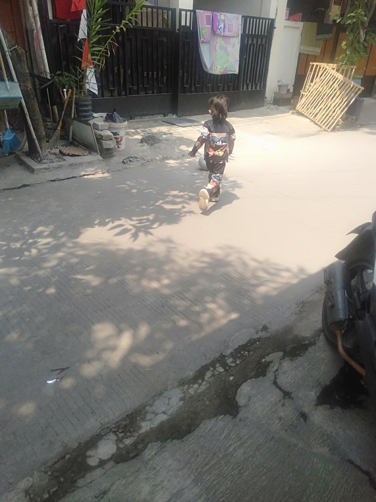 A young child walks away from the camera on a paved street in Ciantra, Indonesia. The child is wearing a long-sleeved shirt and pants with colorful patterns. They are also wearing light-colored shoes. The street is cracked and has some puddles or wet patches. To the left of the child, there is a collection of outdoor items including brooms, a shovel, a bucket, and a plant. In the background, a black metal gate separates the street from a building. A wooden pallet structure is visible to the right of the gate. A motorcycle is parked on the right side of the frame. The scene is illuminated by sunlight, casting shadows of leaves on the ground.