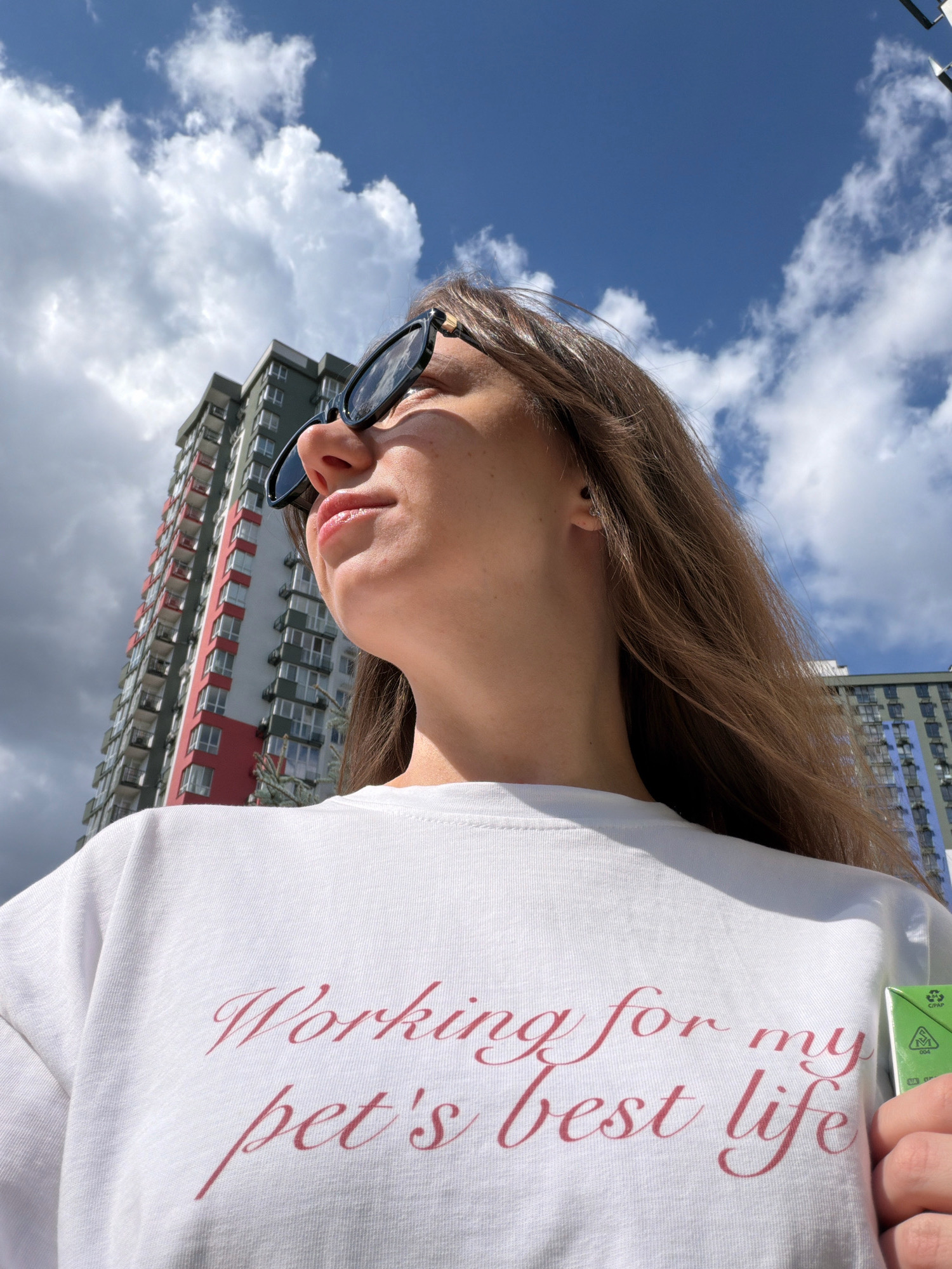 A woman wearing sunglasses and a white t-shirt with the text "Working for my pet's best life" printed on it looks up at the sky. She is standing outdoors with a tall residential building in the background. The sky is blue with white clouds. The building appears to be a modern high-rise, possibly located in a city.