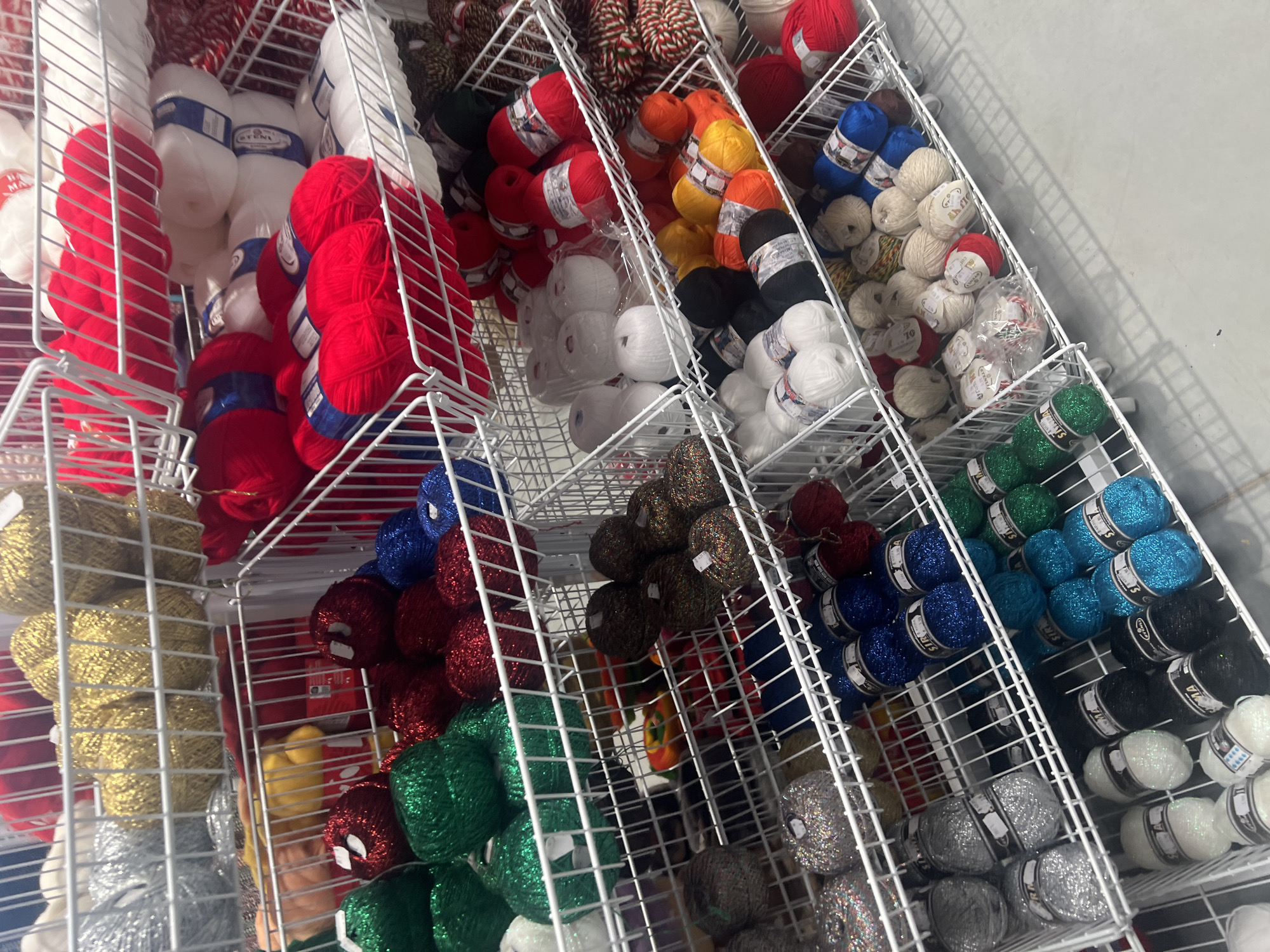 The image shows a retail display of yarn in metal wire baskets. The yarn is in various colors, including white, red, green, blue, gold, silver, black, orange, brown, and multicolored. Some of the yarn has a metallic or glittery finish. The baskets are stacked vertically. There is an off-white wall in the background. It is likely a yarn or craft store in Burgas, Bulgaria, although no external location markers are visible in the image.