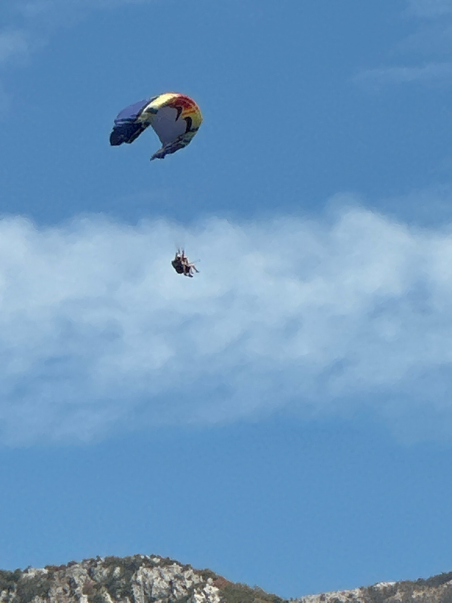 In the city of Boreti, Montenegro, two paragliders are visible against a clear blue sky with scattered white clouds. The larger paraglider, in the upper left portion of the image, is multi-colored with blue, yellow, red, and black accents. A second, smaller paraglider is positioned below and to the right of the first. It appears to be carrying two people who are suspended in a harness. In the lower portion of the image, the rocky, vegetated slopes of a mountain range are visible, suggesting a coastal or mountainous terrain.