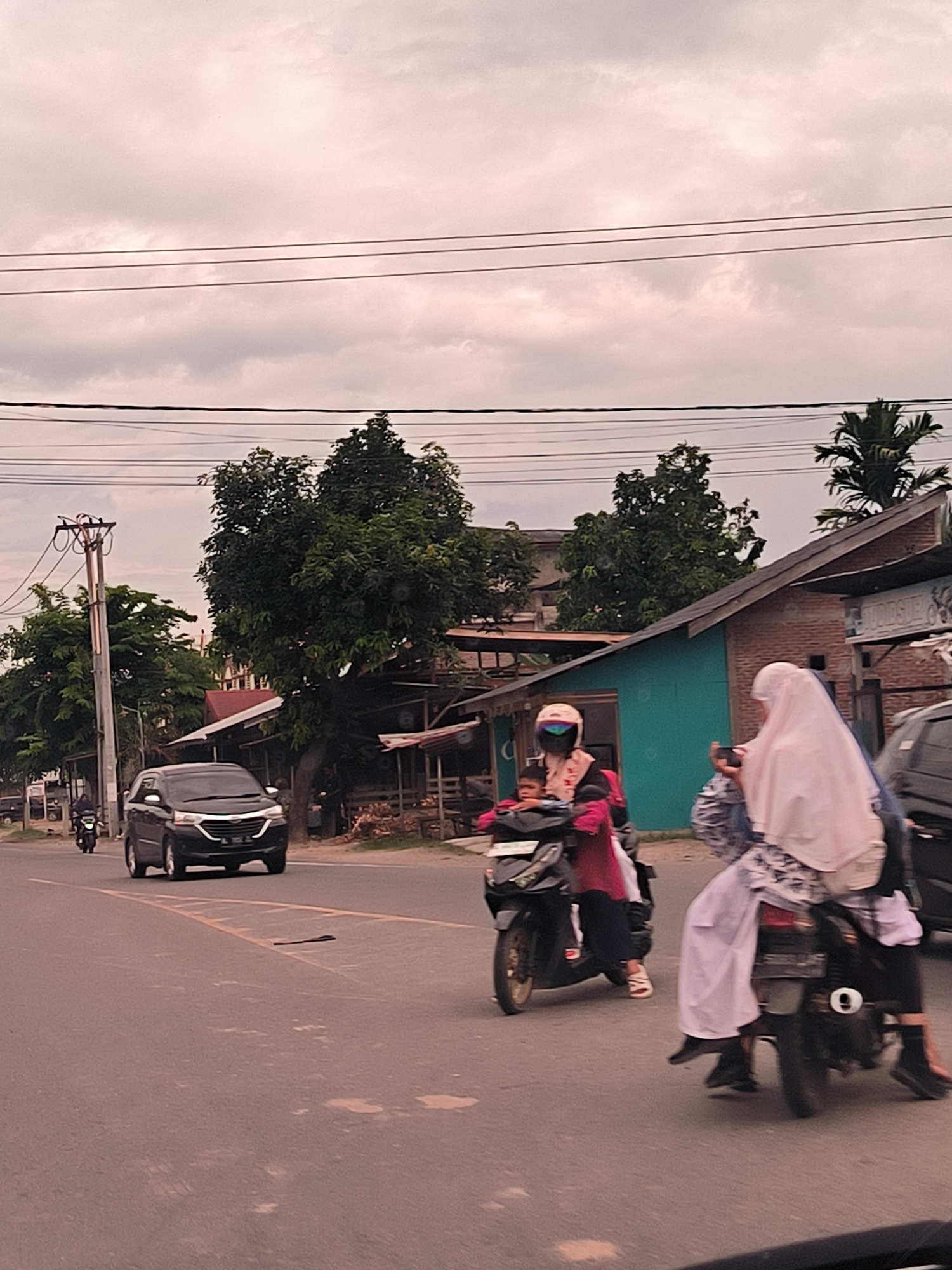 The image depicts a bustling street scene in Payameuneng, Indonesia, under an overcast sky. The atmosphere is diffused and soft, indicating daytime with significant cloud cover.
The primary subjects are vehicles and people. An asphalt road with faded yellow markings leads through the frame. A black multi-purpose car drives towards the viewer in the left lane. On the right side of the road, several motorcycles are in motion. One motorcycle features a driver wearing a pink helmet and visor, with a small child seated in front and another passenger in red behind. Adjacent to this, a black motorcycle carries a driver and at least two passengers. One passenger is a woman wearing a white hijab, holding a smartphone up as if taking a photo or video while riding.
Along both sides of the road, various structures are visible. These include simple residential or commercial buildings, some with teal paint and others with exposed brick, nestled among lush green trees, including palm trees. A prominent wooden utility pole with multiple power lines stands on the left. The scene captures typical daily commute and street activity in a Southeast Asian town. No clearly legible text is visible on signs or vehicles.