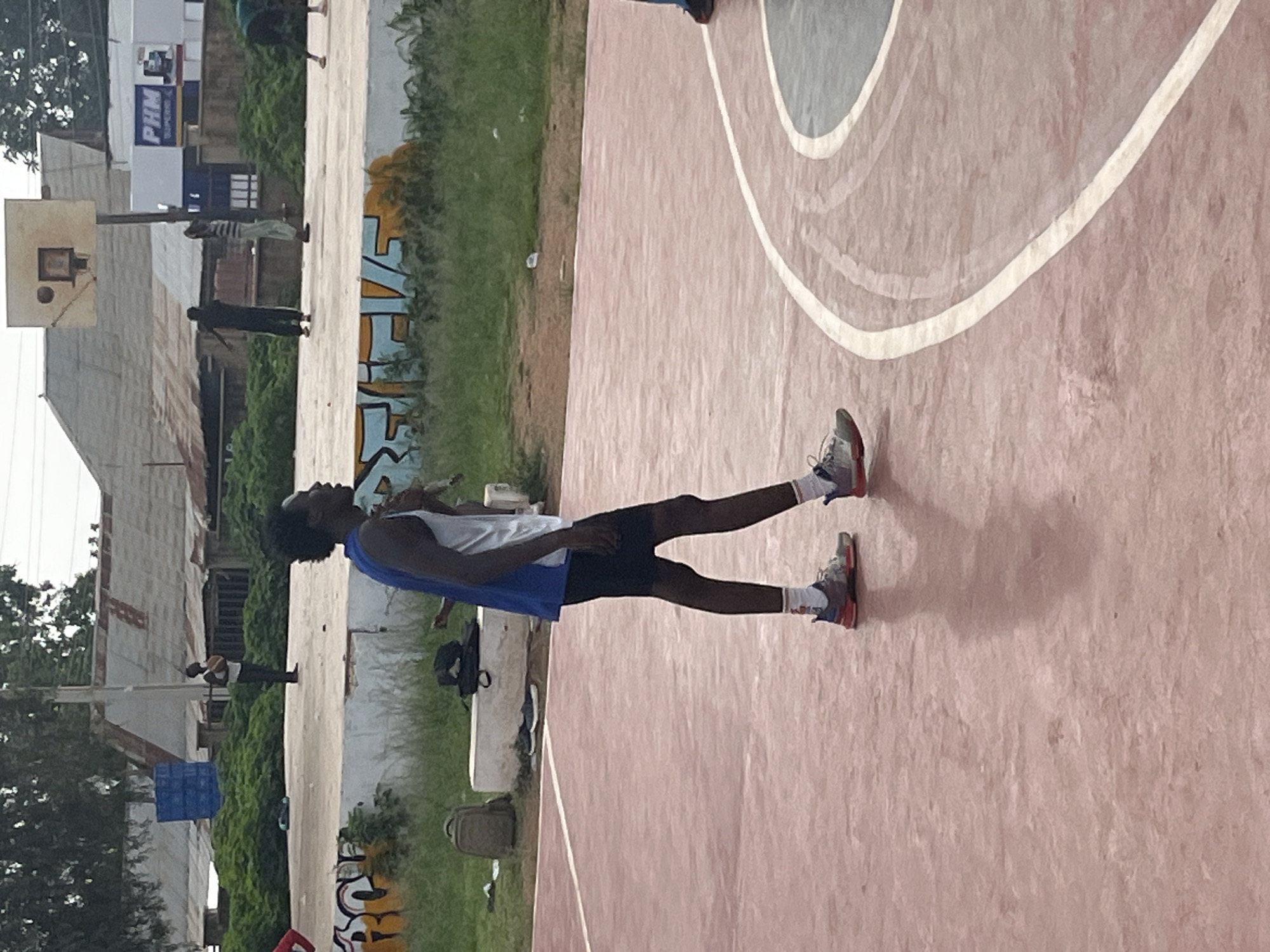 A young Black man, likely in his late teens or early twenties, stands on a pink asphalt basketball court. He is wearing a blue and white tank top and black athletic shorts. His right leg is slightly bent, and his arms are at his sides, suggesting he is either playing basketball or observing. He has short, dark, curly hair and is looking to his left. The court is lined with white paint. In the background, a white concrete bench with a dark backpack and a brown backpack on it is visible. Behind the bench, there are trees and buildings, including structures with corrugated metal roofs, typical of a residential or informal settlement. Further in the background, a basketball hoop is present, and several other individuals are visible in the distance, some near the hoop and others further away, indicating activity on the court. The overall scene suggests a casual outdoor basketball game in a community setting in Jos, Nigeria.