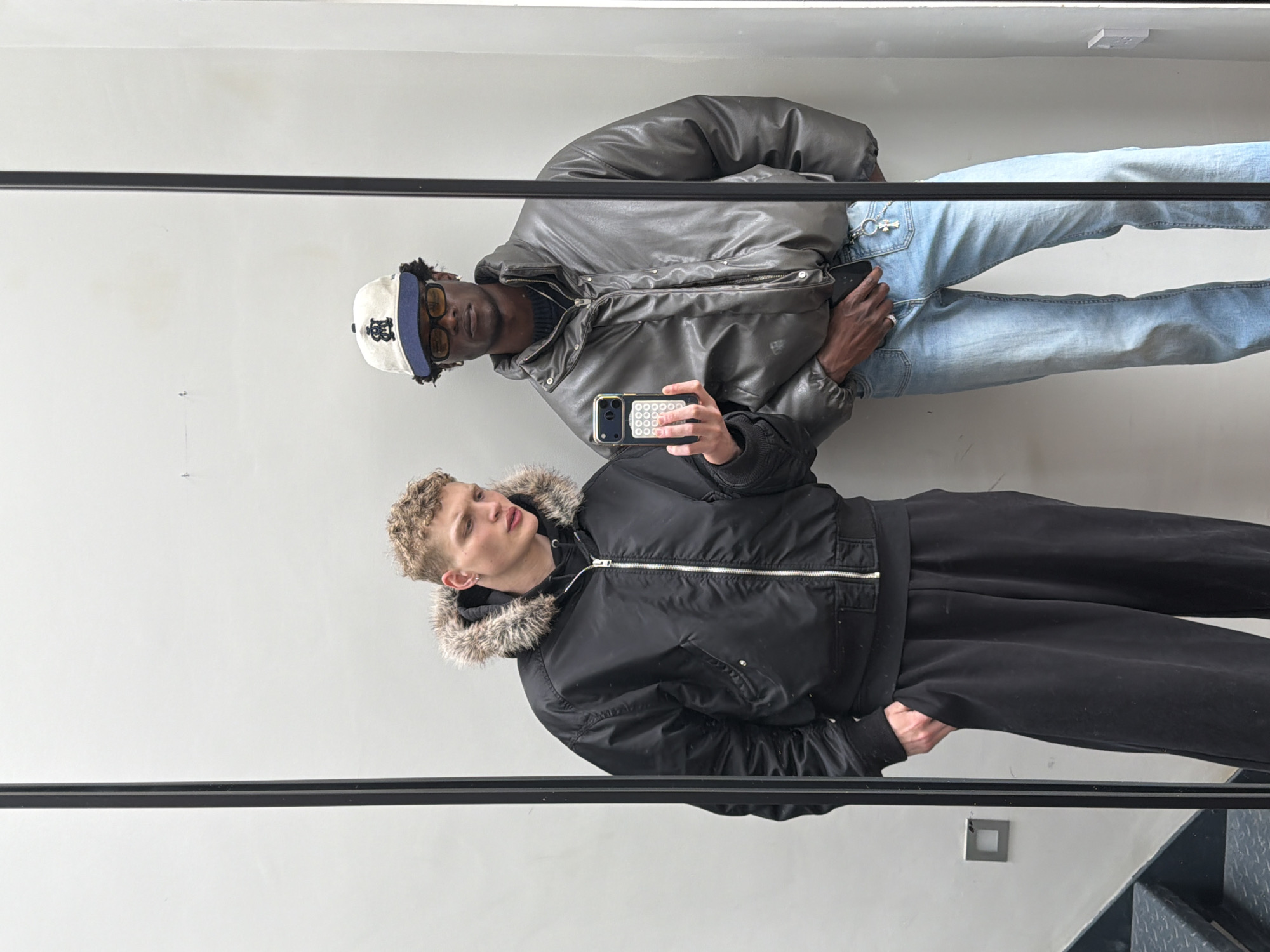 Two adult males are depicted in a mirror selfie taken indoors in London, United Kingdom. The image captures their reflection in a large rectangular mirror with a dark vertical frame down the center.
The subject on the left, with light skin and short curly blonde hair, wears a black bomber jacket with a fur-lined hood, a black hooded sweatshirt underneath, and black wide-leg trousers. He holds a smartphone with a white grid-patterned case in his left hand, aimed at the mirror, while his right hand is in his pocket. He looks downwards and to the right.
The subject on the right, with darker skin, wears a white and navy baseball cap featuring a dark logo, yellow-tinted rectangular glasses, a dark grey puffer jacket, a dark knit sweater, and light blue jeans. A silver chain with a cross charm hangs from his right front belt loop. His left hand is in his pocket, and his right hand rests near his waist. He looks directly forward.
The reflected background consists of a plain white wall. In the lower-left, a dark metal staircase with diamond-plate treads is visible. A square, silver-colored light switch or outlet is mounted on the wall to the left of the mirror's reflection, and a white electrical outlet is visible on the wall to the right.