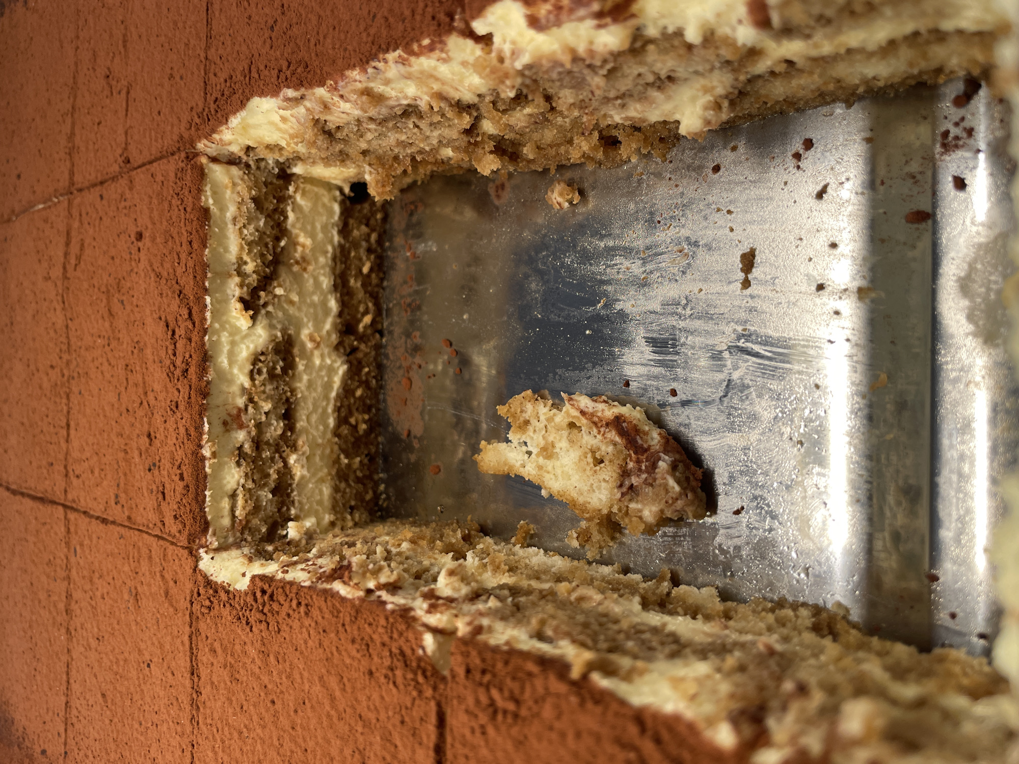 The image depicts a partially eaten Tiramisu cake in a metal baking pan. The cake has layers of ladyfingers, cream, and cocoa powder on top. A piece of the cake is detached and lying in the empty space of the pan. The photo is a close-up shot with an eye-level perspective. The background appears to be a wall or another part of the cake. There is no visible indication of the city of Belgrade, Serbia. No people are visible.