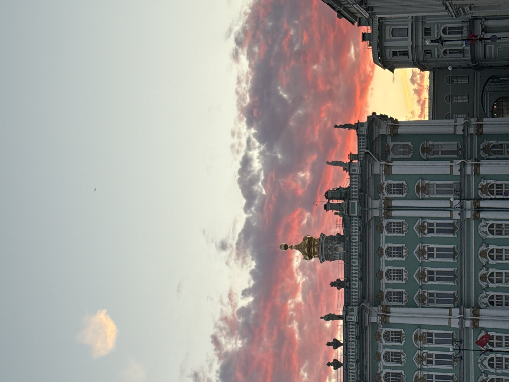 The image shows a view of the State Hermitage Museum in Saint Petersburg, Russia. The sky is a dramatic sunset with vibrant pink and orange clouds. The museum building is a magnificent structure with ornate architecture, painted a distinctive greenish-blue color with white and gold trim. Several windows are visible across the facade. The roofline is adorned with statues and decorative elements, including a prominent golden dome. The scene is captured from a slightly low angle, emphasizing the grandeur of the building against the colorful sky. There is a small cloud formation in the upper left quadrant that is lighter and appears to be illuminated by the sun from behind. In the far upper right corner, a small dark shape, likely an airplane, is visible against the clear sky.