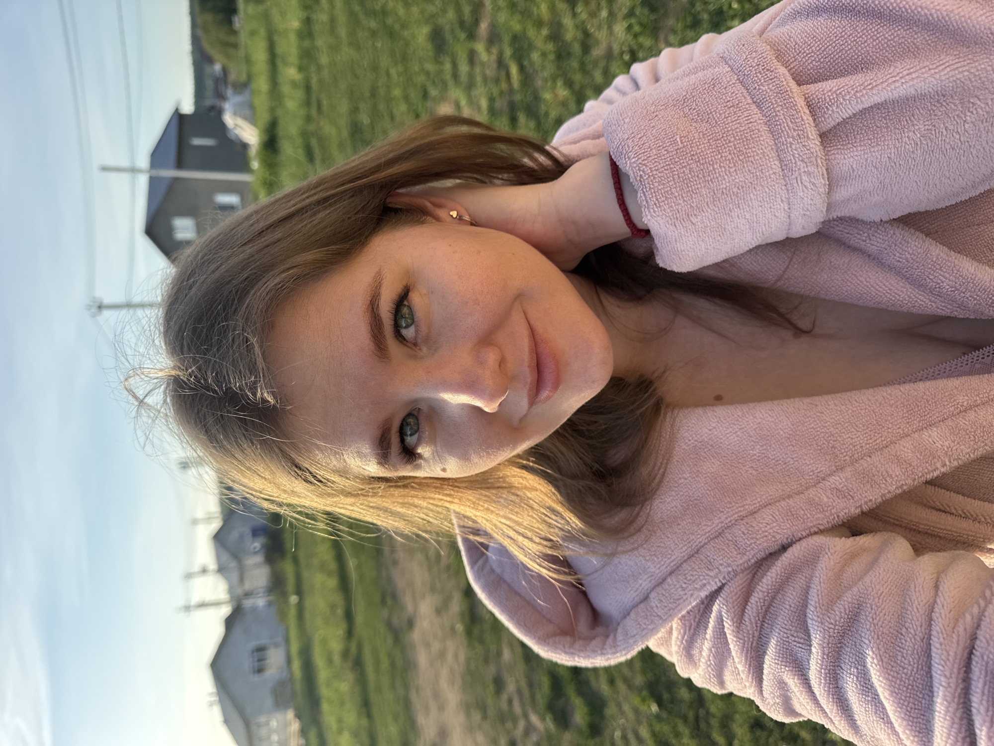 A young woman in a pink bathrobe poses for a selfie outdoors. She is fair-skinned with light brown hair and green eyes. She is smiling slightly, and her left hand is raised to her neck. She is wearing a small gold earring in her left ear and a red bracelet on her left wrist.
The background is a green grassy area with several houses visible in the distance. The houses appear to be located in the city of Ферма, Russia. The sky is blue with some light clouds. The lighting suggests it is either early morning or late afternoon.
