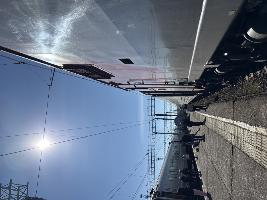 The image displays a train station platform in Kropachyovo, Russia, illuminated by direct sunlight under a clear, bright blue sky. Overhead, a complex network of electrical catenary lines and supporting gantries spans across multiple railway tracks.
On the immediate right, a large train car, presenting a two-tone metallic silver-grey and maroon-red exterior, is stationary. Its reflective metallic surface exhibits significant sun glare and visible circular polishing marks. Multiple vents and access hatches are discernable along its side.
To the left, on an adjacent track, another passenger train, characterized by its silver-grey color and numerous windows, is also stationary.
Between these two trains, on the concrete platform, approximately 10-15 individuals are observed. They are dressed in casual attire and are largely standing in small groups or individually. One person on the far left wears light pink pants. In the central foreground, near a cluster of people, items resembling luggage or personal belongings are situated on the platform. A solitary individual stands near the right-side train, facing away from the camera, possibly near an open doorway or car coupling. The platform surface is concrete, featuring tactile paving strips.