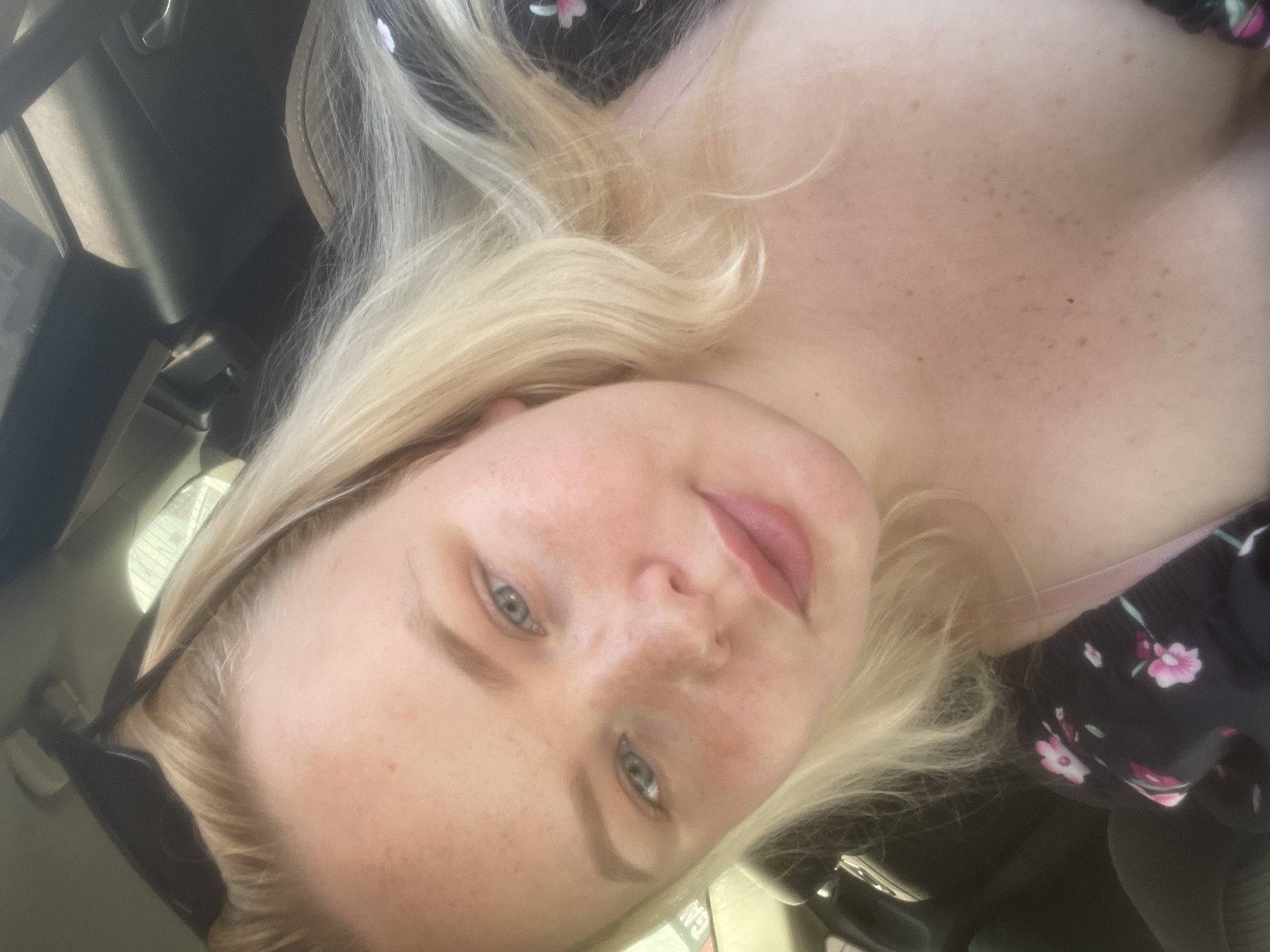 A close-up, selfie-style shot shows a young Caucasian woman with blonde hair and fair skin, appearing to be in a car. She is looking at the camera, with her head tilted slightly. Her eyes are light blue, and she has light makeup on. She is wearing a dark floral print top with pink flowers. The background is dark and blurred, suggesting the interior of a vehicle. The image is well-lit, with natural light illuminating her face. No specific landmarks or elements within the image suggest a location in Samara, Russia, or any other city.