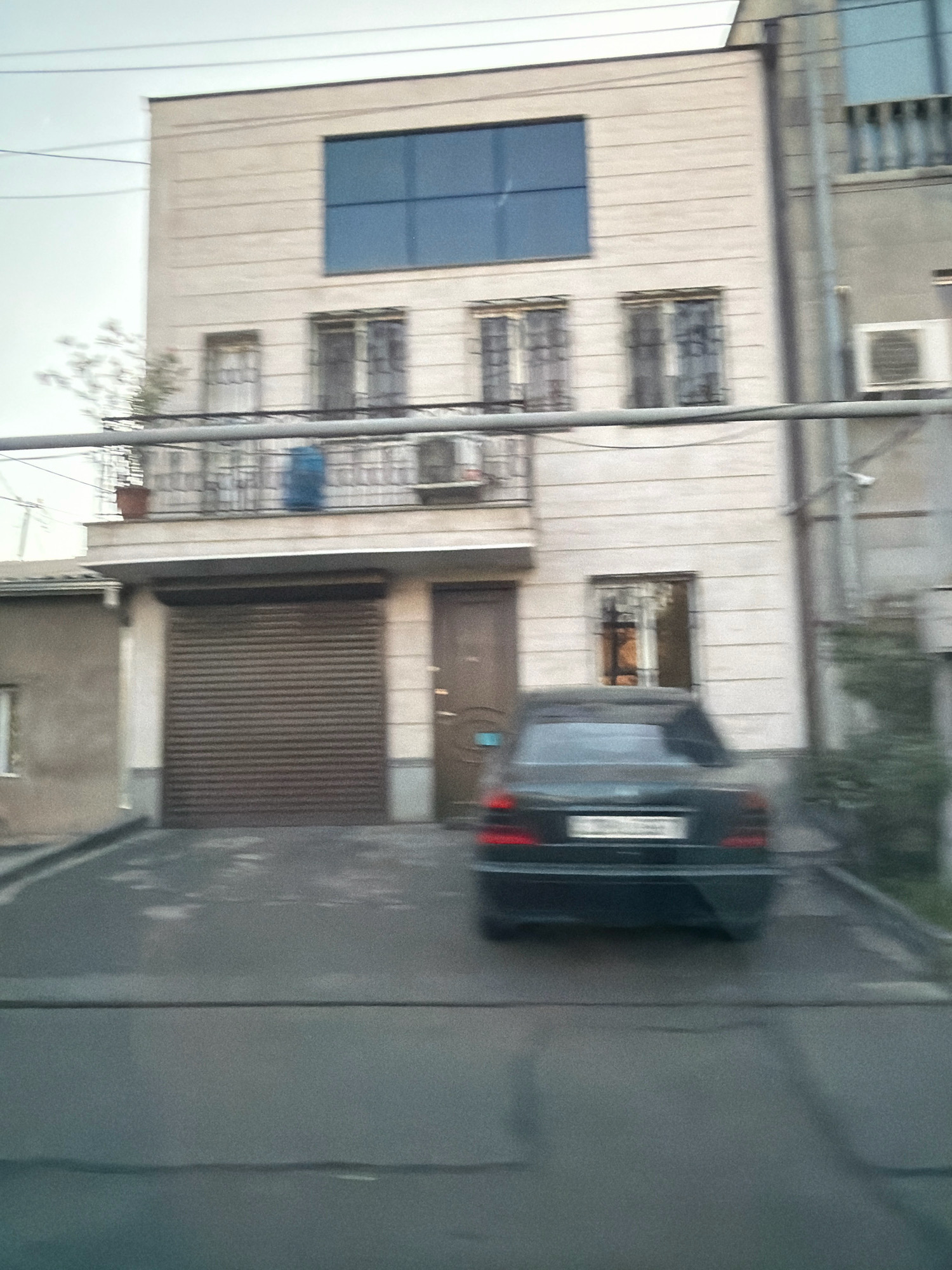 The image depicts a street scene in Yerevan, Armenia, featuring a multi-story building with a light-colored facade. The building has several windows, some with metal grates. A large, dark-colored garage door is visible on the ground floor of the building. A dark sedan is parked in front of the garage. A balcony with a railing is present on the second floor. Utility lines are visible running horizontally across the top of the image. The surrounding environment appears to be an urban setting with a slightly overcast sky.