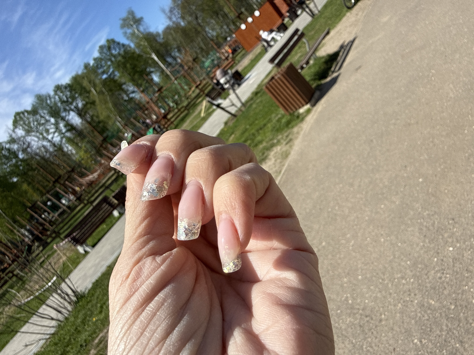 A person's hand with decorated nails is in focus, with the background slightly blurred. The nails have a French manicure style with silver glitter on the tips. The skin on the hand is fair, with visible wrinkles and creases. The background appears to be an outdoor park setting, with trees, green grass, benches, and a walking path. The sky is visible in the upper left corner, indicating a sunny day. Based on location metadata, the park is in Заволжье, Russia.