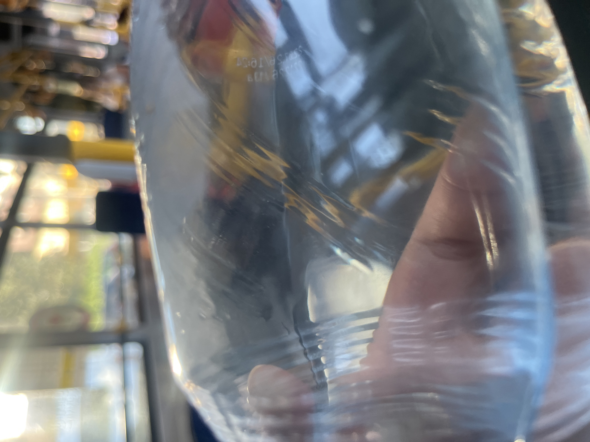 The image shows a close-up view of a transparent plastic bag containing a liquid, likely water, with a hand holding it. The background is blurred, suggesting an indoor environment with light filtering through windows, possibly a public transportation vehicle like a bus or train, given the presence of seats and poles visible in the bokeh effect. The scene is captured in a city, but the specific location within Bishkek, Kyrgyzstan, cannot be determined from the visual information alone. The lighting suggests it is daytime.
