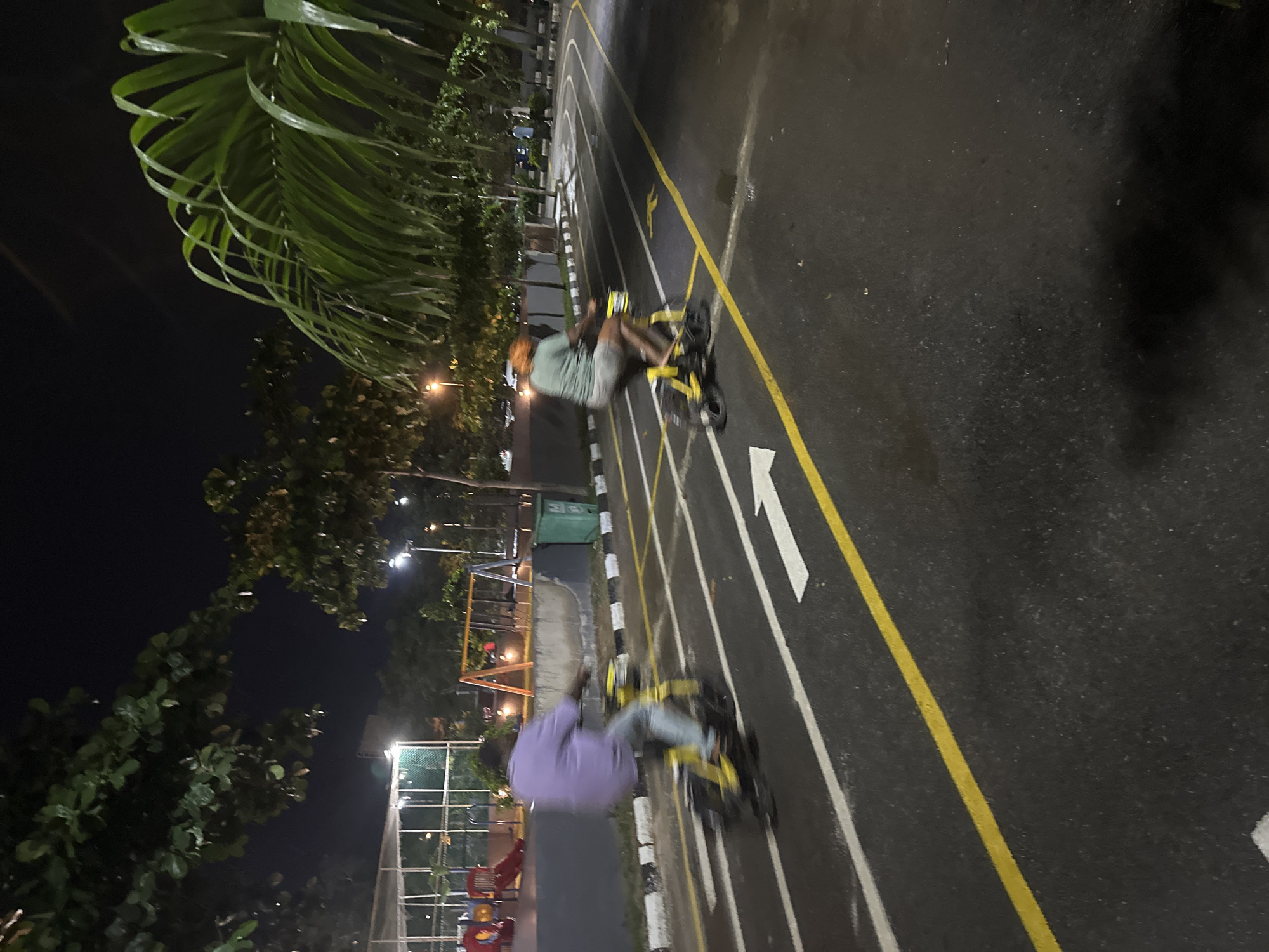 Two individuals are riding yellow bicycles on a paved, dark asphalt path at night in what appears to be a park or recreational area. The path is marked with white and yellow lines, indicating lanes and directional arrows. The person on the left is wearing a purple shirt and blue jeans, while the person on the right is wearing a greenish-blue t-shirt and shorts. Both are in motion, causing a slight blur in the image. In the background, there is a playground with slides and swings, illuminated by overhead lights. Green trees and foliage are visible around the perimeter of the path and playground. The scene is consistent with an outdoor public space in Abuja, Nigeria, likely a park with cycling or running tracks and a playground.
