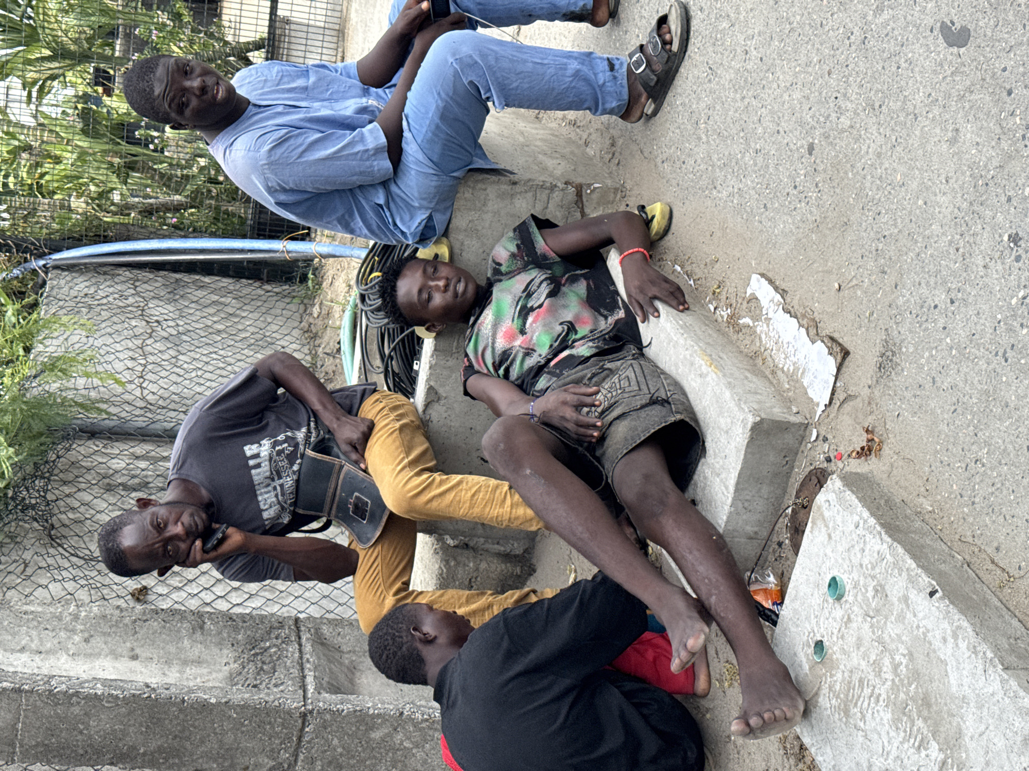 Four young Black men are depicted resting outdoors on concrete structures in what appears to be Lekki, Nigeria, under bright daylight conditions.
In the upper left, a man wearing a light blue tunic and blue trousers sits on a concrete step, looking towards the camera with a slight smile. He has dark sandals on his feet. Directly below him, another man with dreadlocks lies barefoot on a lower concrete slab, seemingly resting or asleep. He wears a multi-colored t-shirt with a green, black, and red pattern, and grey shorts, with a red bracelet on his wrist.
To the left of the lying man, a third man is seated on concrete steps, holding a mobile phone to his ear and appearing to be on a call. He wears a dark grey t-shirt with "TENNESSEE" visible on it, and mustard-yellow trousers. A black pouch or bag is slung over his shoulder. A fourth man, wearing a black top and red trousers, is partially visible in the bottom right, also resting on the concrete with his bare feet.
The setting includes concrete steps and blocks, a metal mesh fence behind the subjects, and green foliage in the background. The ground is a mix of concrete and dirt, with various items like a blue pipe, electrical cables, and some debris scattered around. The overall scene suggests a casual gathering or resting period in a public or semi-public outdoor area.
