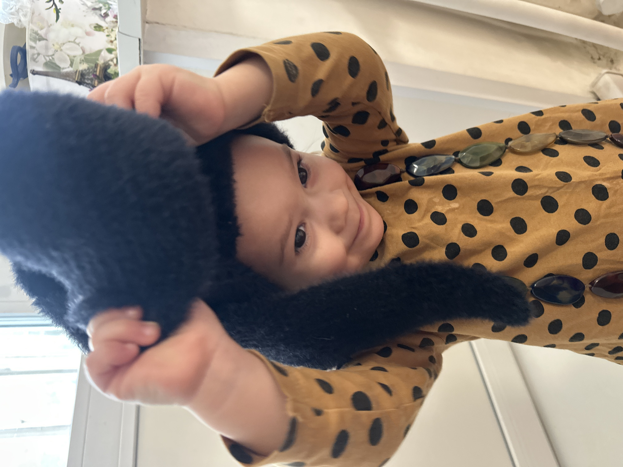 This image, captured in Uva, Russia, features a close-up, rotated portrait of a young child, likely a baby or toddler. The child's face is central, looking directly at the camera with a gentle, curious expression, bright eyes, and a slight, contented smile.
The child is wearing a plush, black, fuzzy hat that covers their ears and frames their face. They are dressed in a mustard yellow or light brown long-sleeved shirt patterned with distinct black polka dots. Around the neckline of the shirt, a decorative element is visible, composed of several large, oval-shaped, translucent beads in muted shades of green, brown, purple, and blue. The child's small hands are visible at the top and bottom of the frame, grasping the fuzzy hat.
The background is softly blurred, suggesting an indoor setting. A bright area on the left hints at a window or a strong light source, with faint details that could be white curtains or artificial foliage. White wall sections or door frames are discernible on the right and bottom edges of the background. The natural light indicates it is daytime. No text is visible in the image. The composition conveys an intimate and playful moment.