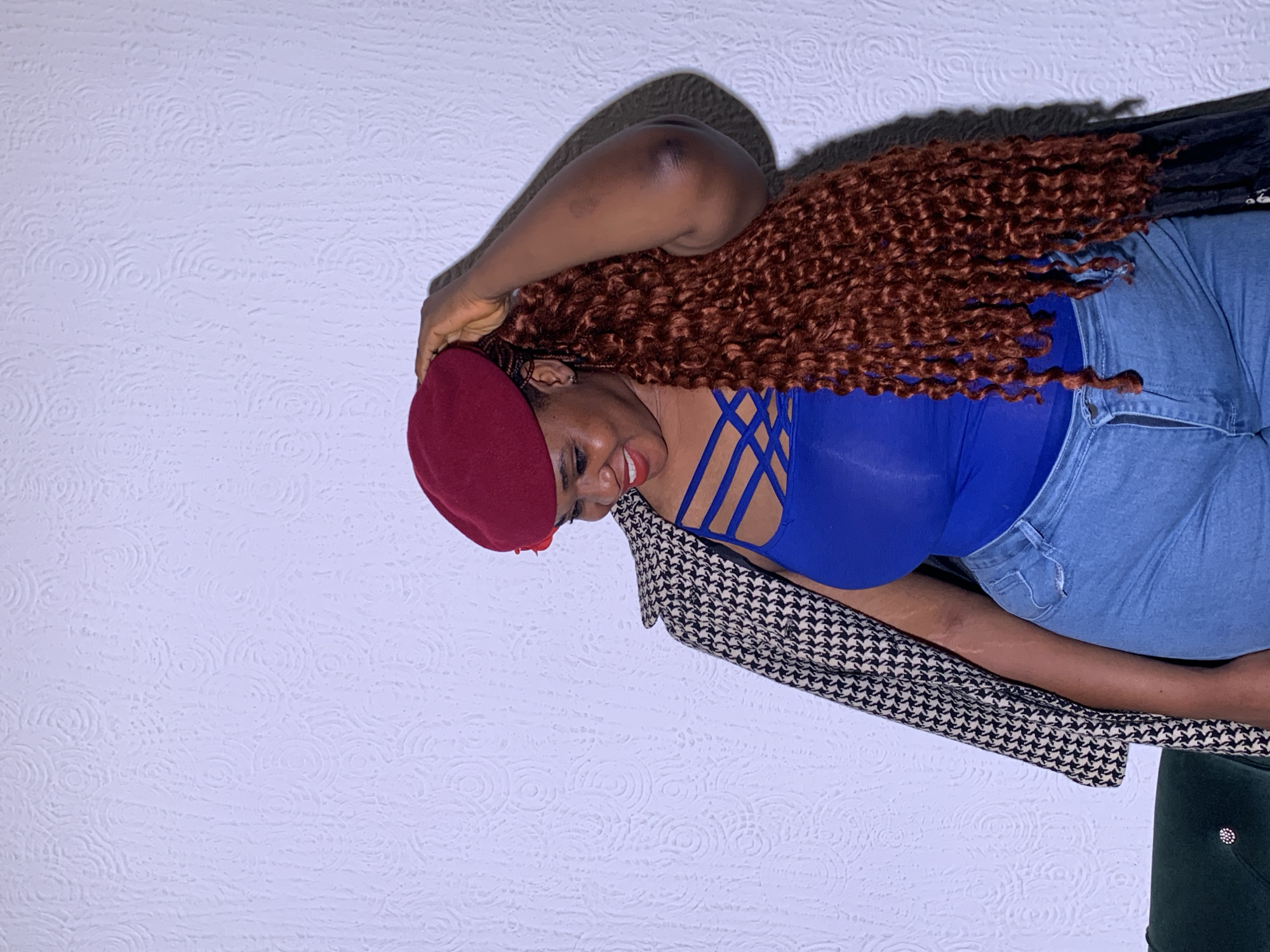 An adult woman is captured in a medium close-up, leaning sideways against a textured wall. She is smiling broadly with red lipstick visible, and her head is tilted. She wears a dark red beret and has long, reddish-brown curly hair. Her attire includes a bright blue top with criss-cross strap details over the bust, paired with light blue denim jeans. A black and white patterned garment, possibly a jacket or scarf, with a houndstooth-like design, is draped over her arm. Her right arm is raised, with her hand near her beret.
The background features a light-colored wall with a repeating embossed circular or swirling pattern. Shadows are cast on the wall behind her, suggesting indoor lighting. A dark green, possibly velvet, piece of furniture is partially visible in the bottom right corner. The overall mood is cheerful due to the woman's smile. No visible text is present. The context provided indicates the location is Barakin Akawo, Nigeria.
