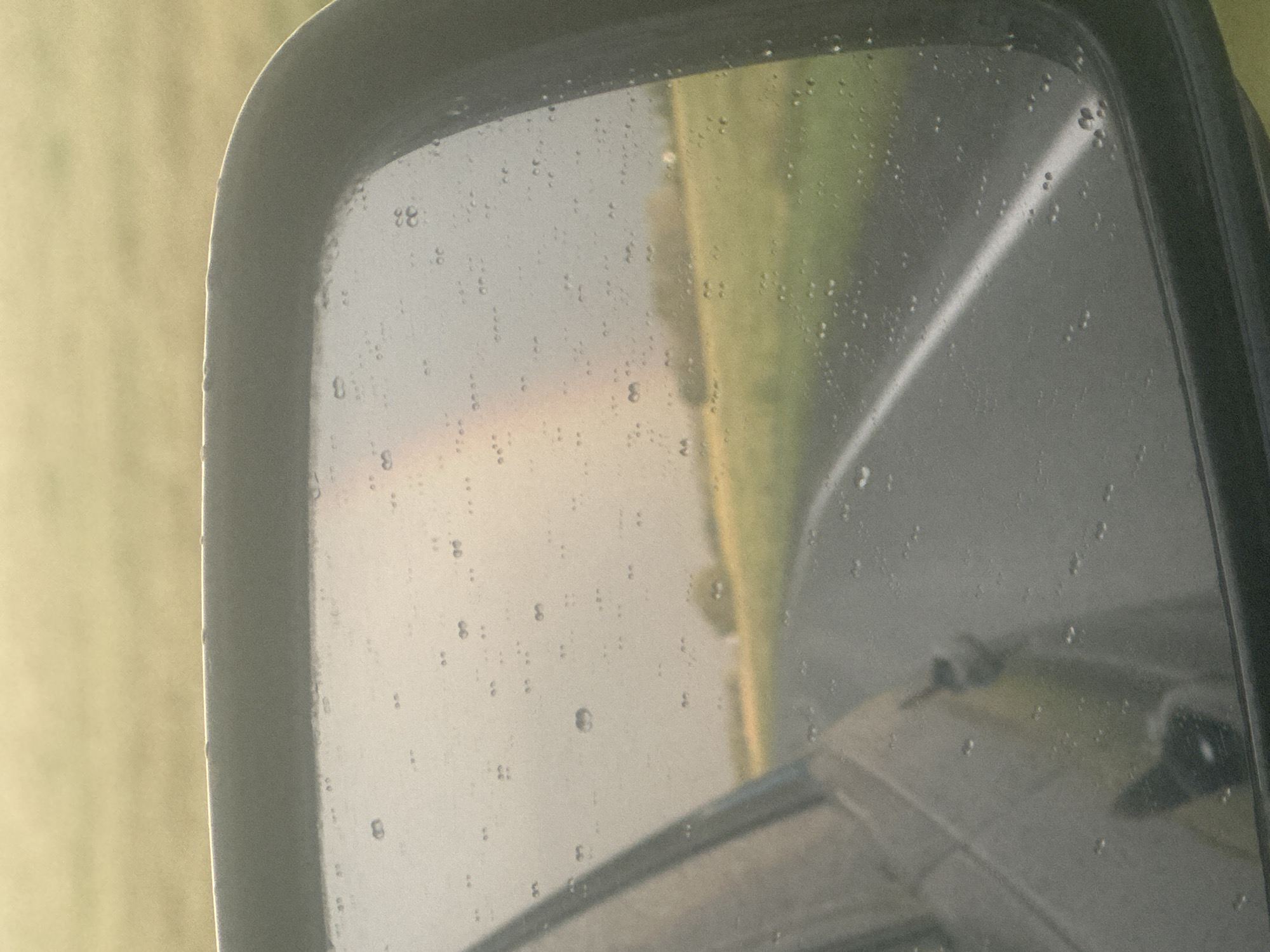 The image shows a rearview mirror of a vehicle with raindrops on its surface. Through the mirror, a rural landscape is visible, with a road, green fields, and a faint rainbow in the sky. The reflection in the mirror shows the interior of the car, including a portion of the driver's side door and the window. The weather appears to be overcast and possibly rainy, given the presence of raindrops and the subdued lighting. The location in Pastukhova, Russia, is not evident from the image itself.