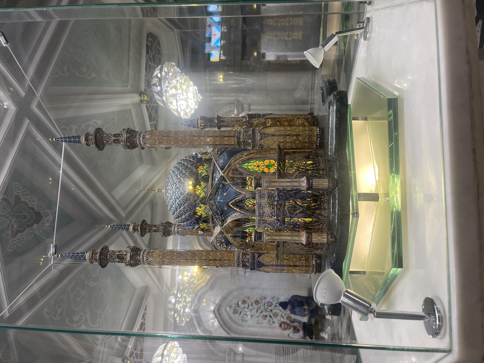 A detailed model of the Kul Sharif Mosque is encased in a glass display. The model features a central dome, multiple minarets, and intricate ornamentation. The structure appears to be made of metal and glass. It is illuminated by two small spotlights attached to the display case. The display is located inside the actual Kul Sharif Mosque in Kazan, Russia. Reflections in the glass case show elements of the mosque's interior, including ceiling decorations, light fixtures, and blurred figures of visitors. The overall environment suggests a museum-like setting within a place of worship.