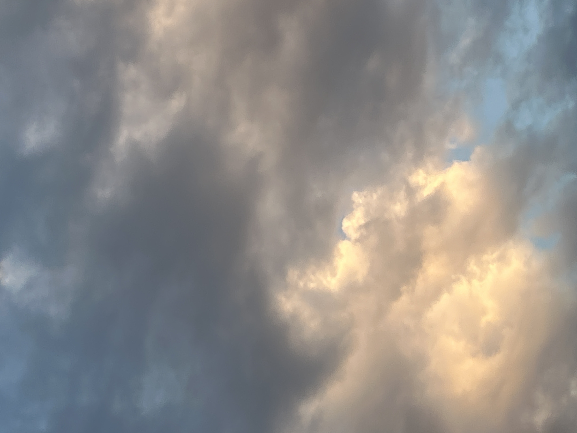 This is a photograph of a cloudy sky. The clouds are a mix of dark grays and lighter, warmer tones, suggesting they are illuminated by sunlight. There is a small patch of blue sky visible towards the upper right. The lighting implies it is either sunrise or sunset. The overall scene is one of natural beauty, with the clouds forming interesting shapes and textures. There is no text visible in the image.