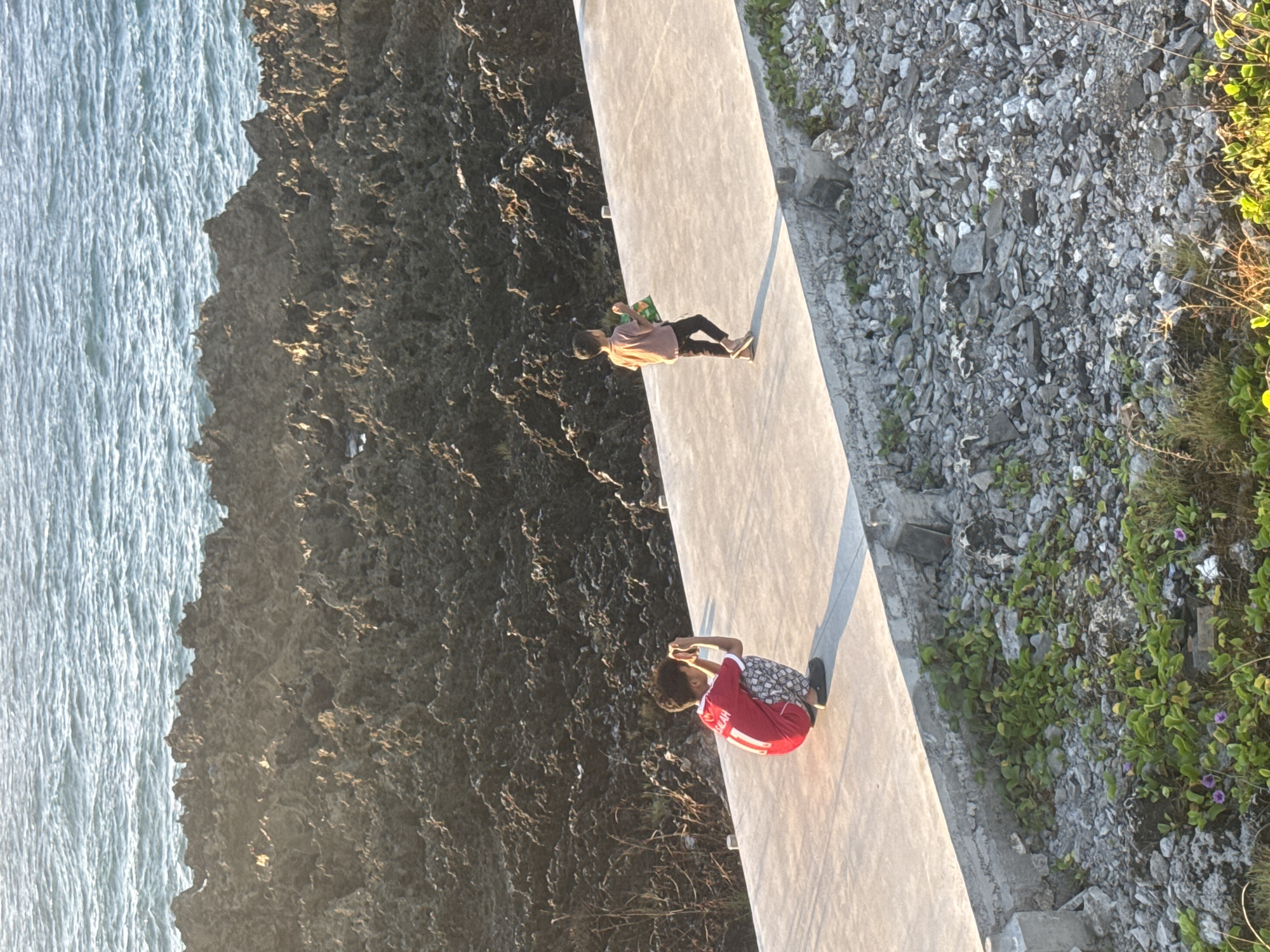 The image captures a sunlit coastal scene in Kuta Selatan, Indonesia, during what appears to be the late afternoon, indicated by the warm light and long shadows. The photograph is oriented as if looking down from an elevated position.
A light-colored concrete pathway or wall stretches horizontally across the midground. To the left of this pathway, a rugged, dark rocky coastline borders the ocean, which shows a textured surface from gentle waves. To the right of the pathway, the ground is covered with grey, irregular rocks and interspersed with green vegetation, including small bushes and plants bearing delicate purple flowers.
Two individuals are visible on the pathway. Further away, one person stands, wearing a light-colored t-shirt and dark pants, holding a green object, possibly a bag or box, in their hands. Closer to the viewer, another person is crouched down, engrossed in a smartphone or similar small device held in their hands. This individual wears a red t-shirt with "SALAH" and the number "11" prominently displayed on the back, indicative of a football jersey, along with patterned shorts. They have curly hair.
The overall atmosphere suggests a leisurely moment by the sea, with both individuals engaged in their personal activities amidst the scenic coastal environment.