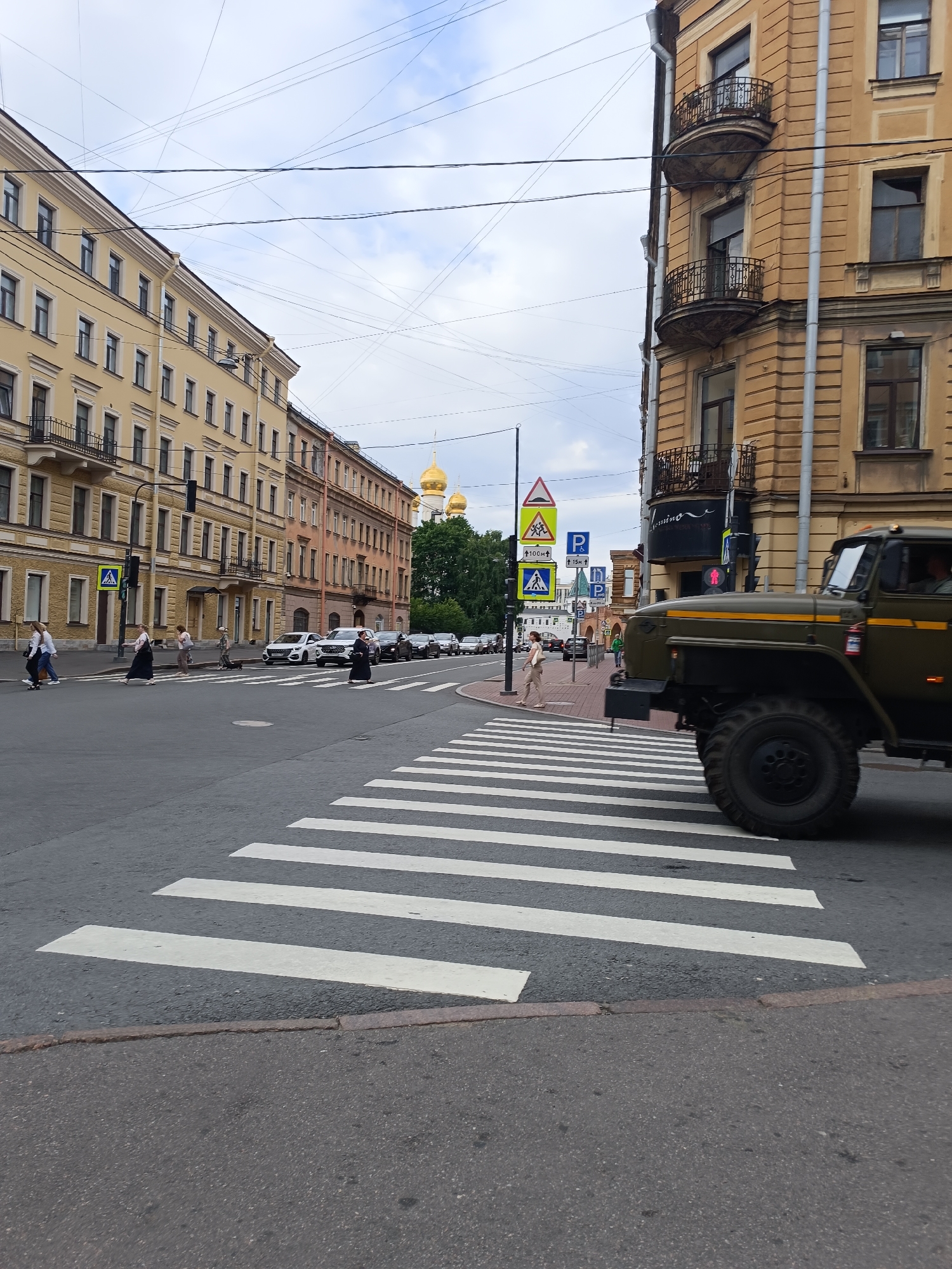 The image shows a street scene in Saint Petersburg, Russia, featuring a zebra crossing in the foreground. To the right, a large, olive-drab military truck is partially visible, its front wheel prominently displayed on the zebra crossing. In the background, multi-story buildings with traditional St. Petersburg architecture line the street. The building on the right is ochre-colored with decorative balconies, while the buildings on the left are lighter yellow. Golden domes of a church are visible in the distance between buildings, indicating a prominent religious site. Several pedestrians are crossing the street at the zebra crossing, some dressed in dark clothing. Traffic lights and various road signs are also present. The sky is overcast with clouds. The overall impression is a typical urban environment in Saint Petersburg, with a mix of historical architecture and modern city life.
