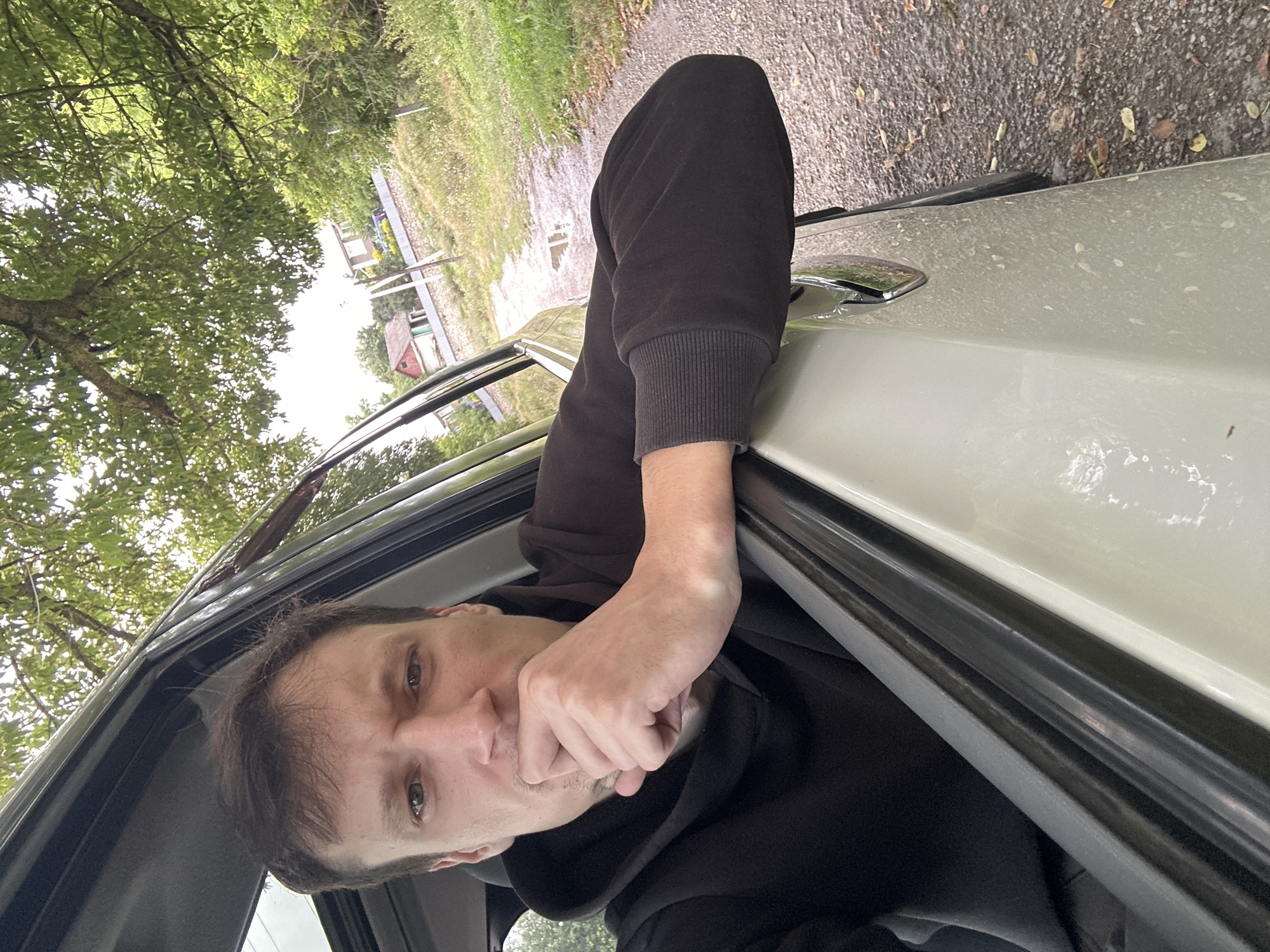 A man is taking a selfie from inside a vehicle with his right arm resting on the open car door and his hand near his mouth. He is wearing a black hoodie. The vehicle is parked on the side of a rural, gravel road with grass and trees on either side. In the background, there are houses and a fence. The scene appears to be in a rural area.