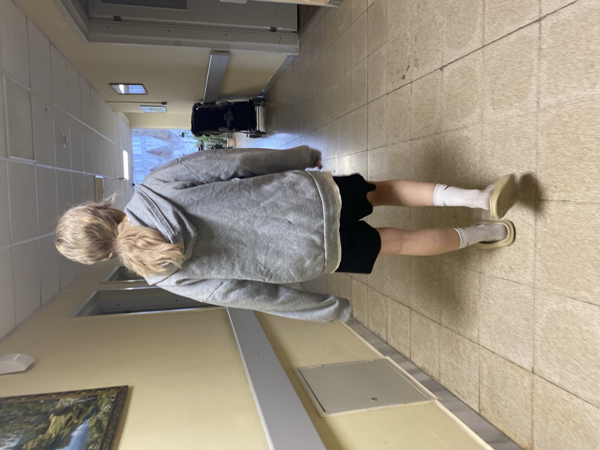 A person is standing in a hallway. They are wearing a gray hoodie, black shorts, white socks, and light-colored sandals. The hallway has tile floors and light yellow walls. A wheelchair is visible further down the hallway. A painting is hanging on the wall near the person. The location is Марковский, Russia.