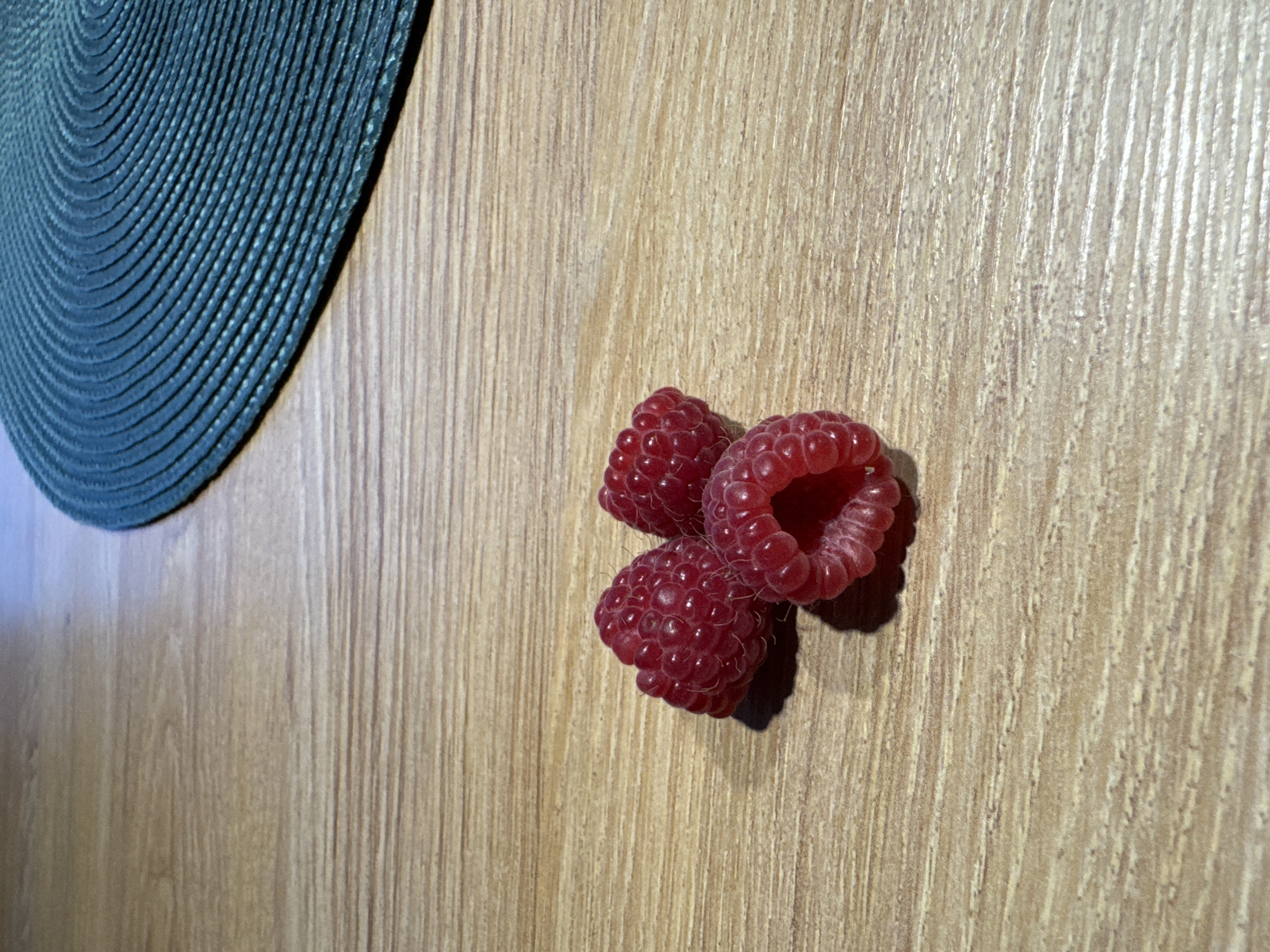 A close-up, overhead shot shows three ripe raspberries clustered together on a light-brown wooden surface. The raspberries are a deep red, with visible segments and a slightly fuzzy texture. One raspberry is partially hollowed out, suggesting it has been eaten. To the left of the raspberries, a portion of a dark teal placemat is visible, its texture appearing to be woven or ribbed in a concentric pattern. The wooden surface exhibits a prominent grain running vertically. Sunlight appears to be casting a shadow from the raspberries onto the wood, suggesting the light source is from the right and slightly above. The overall impression is of a still life, focusing on the textures and colors of the fruit and its surroundings. No people are visible, and no text is present in the image. The setting appears to be indoors, on a table or countertop.