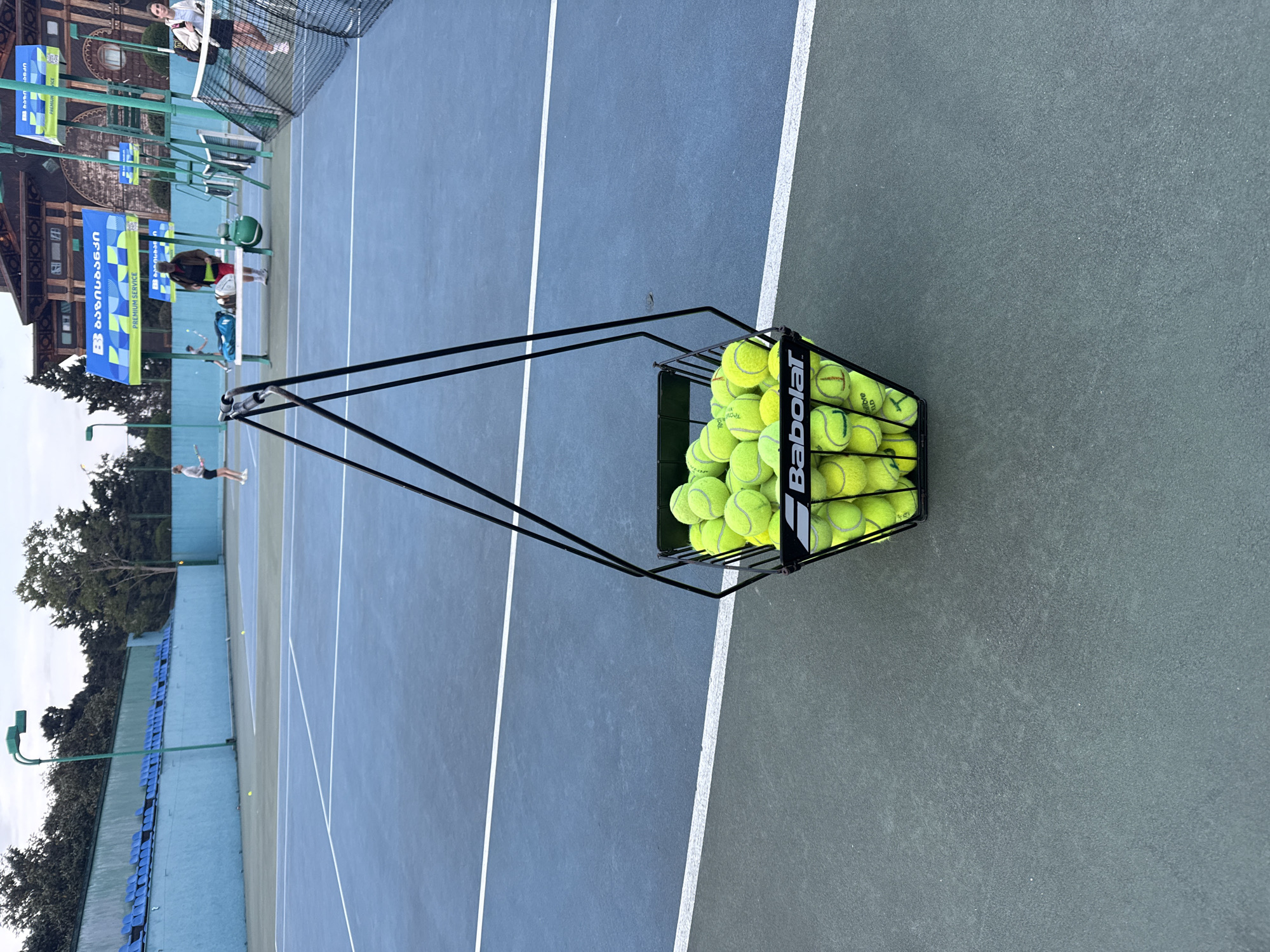The image depicts a tennis court in Batumi, Georgia. A Babolat ball hopper filled with tennis balls sits in the foreground near the right edge of the frame. The tennis court is painted blue and green, with white lines marking the boundaries. In the background, several people are playing tennis, and spectators can be seen near the net. A building with traditional Georgian architectural elements is visible behind the tennis court. Banners written in Georgian line the back fence. Blue plastic chairs make up a section of stadium seating along the side of the court.
