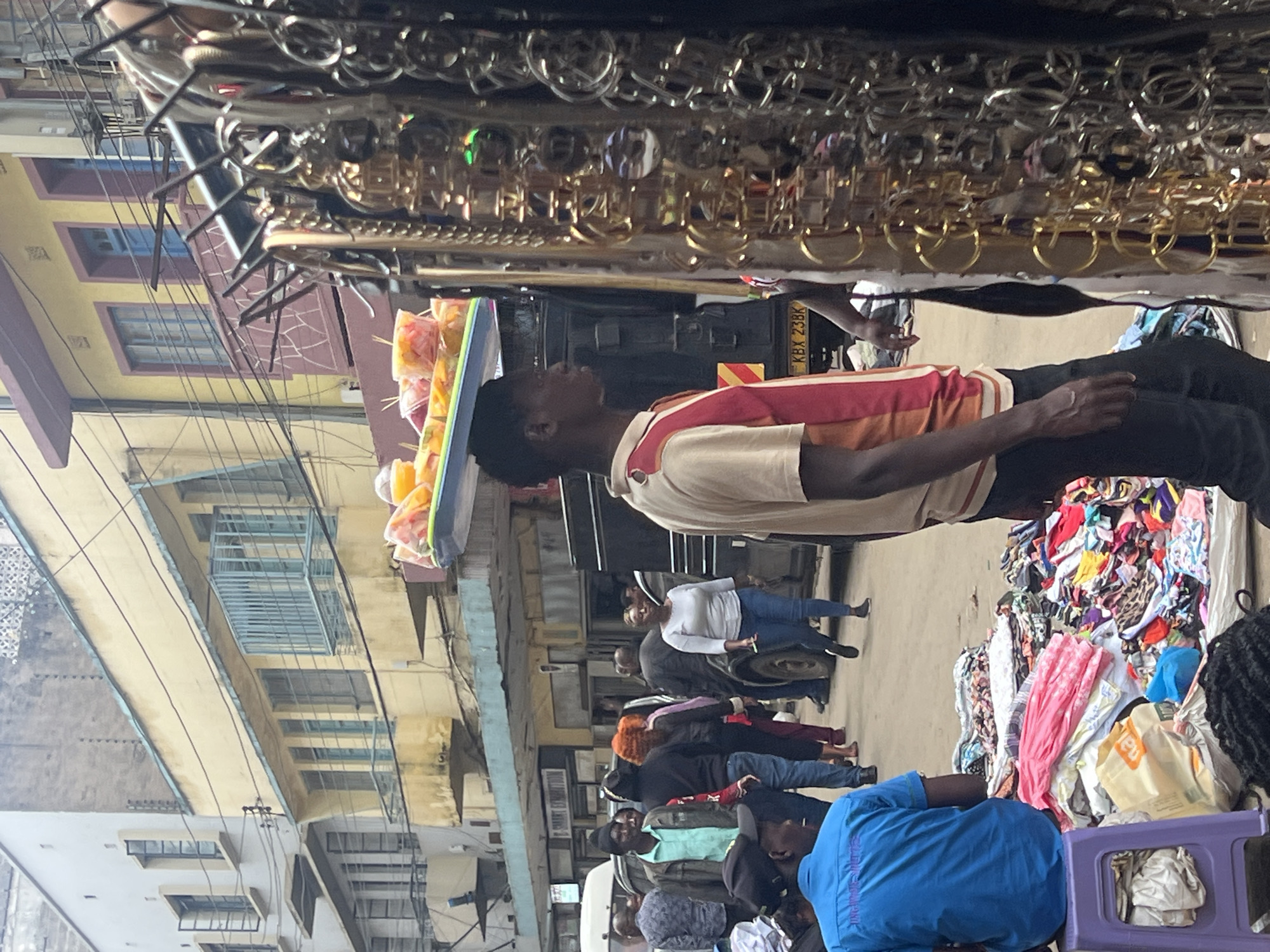 The image captures a bustling street scene in Nairobi, Kenya, during a bright, sunny daytime. The primary subject in the mid-foreground is a young man, dressed in a red, white, and orange striped short-sleeved shirt and dark trousers, skillfully balancing a blue tray of plastic-wrapped, chopped fruits on his head, likely selling them. He looks forward, navigating the crowded environment.
Immediately above him, in the top part of the frame, is a dense display of metallic objects, possibly jewelry such as necklaces, earrings, or decorative keyrings, hanging from a market stall rack. These items vary in color, appearing both gold and silver-toned.
Behind the fruit vendor, a black utility vehicle or van is visible, featuring a Kenyan license plate "KBX 23BK". The street is filled with other people engaged in various activities; a woman in a white top and blue jeans is seen, along with several other pedestrians and shoppers in casual wear. On the right side, a large, colorful stack of clothes indicates a textile stall, with a partially visible yellow bag bearing the text "DAL" resting nearby. In the lower right, a person in a blue shirt is partially seen, possibly sitting on a purple plastic stool.
The background is dominated by multi-story urban buildings, predominantly in shades of light yellow and beige, featuring blue-framed windows and balconies with metal railings. Overhead, numerous electrical wires crisscross between the structures. The scene depicts a lively, commercial atmosphere characteristic of an urban market.