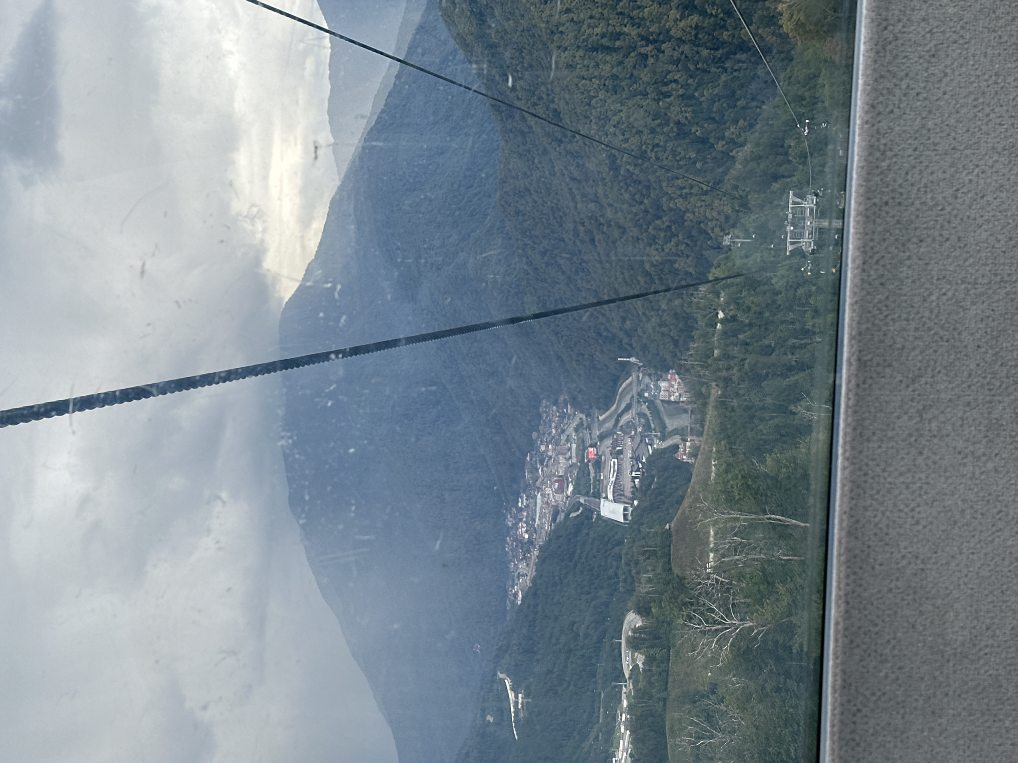 The image shows a view of the mountain resort town of Estosadok, Russia, as seen from a cable car. The town is nestled in a valley surrounded by lush green mountains. A river runs through the valley, and various buildings, including hotels and ski lifts, are visible. The sky is overcast, with thick clouds obscuring the sun. The cable car's support cables are prominently featured in the foreground, with a ski lift station visible further down the slope. The overall impression is one of a scenic and serene mountain landscape.