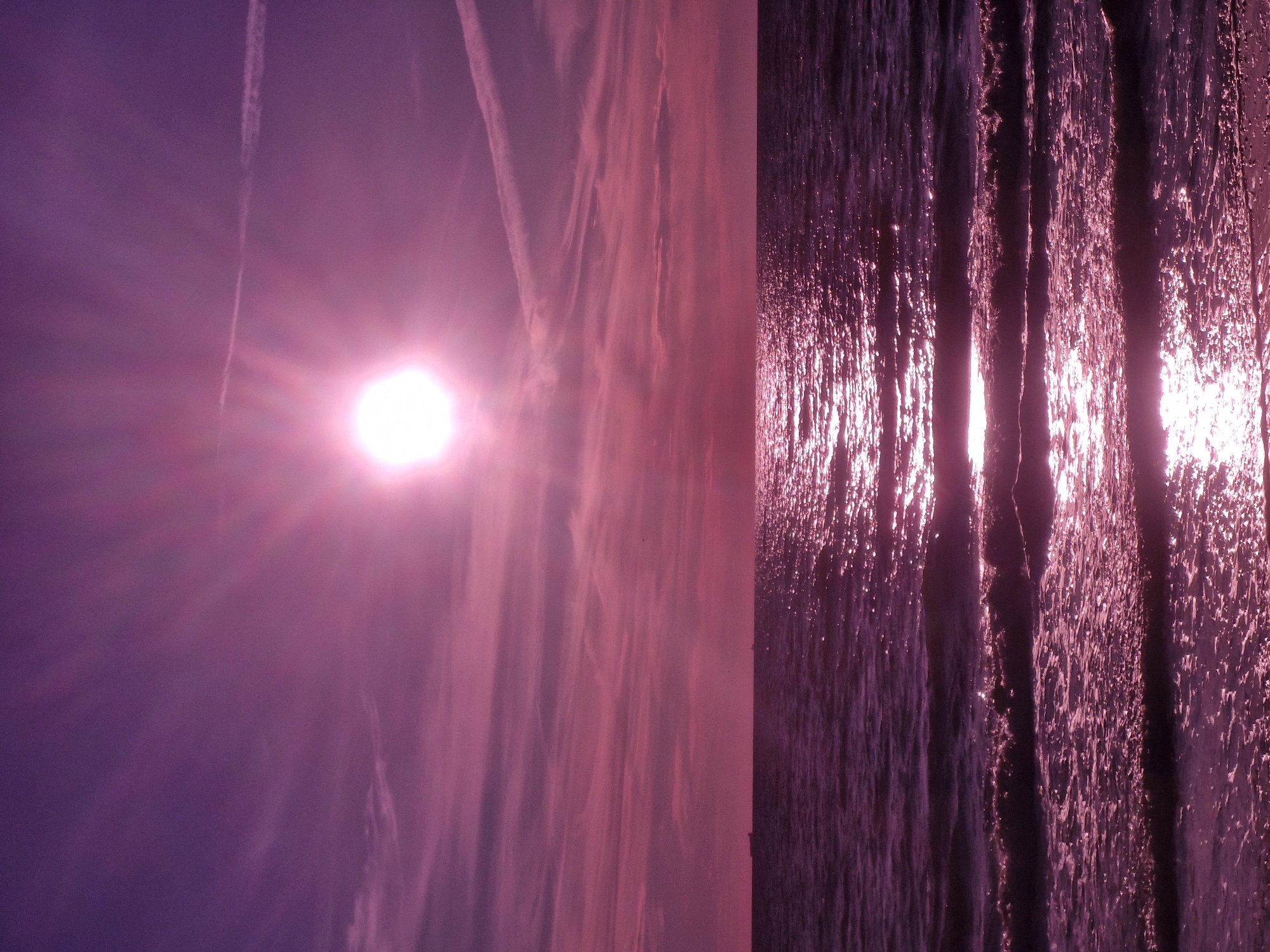 The image depicts a serene ocean scene bathed in a vibrant purple hue, likely due to a filter or the time of day. The sun, a brilliant white orb with visible rays, dominates the upper portion of the sky, casting a shimmering reflection across the water's surface. Wispy clouds stretch across the sky, some forming thin, elongated trails. The sea itself is calm, with gentle waves lapping at the shore, creating patterns of light and shadow. In the distance, two dark shapes, possibly ships, are visible on the horizon. The foreground shows the edge of a beach where the water meets the sand, with foamy wavelets. The overall atmosphere is tranquil and visually striking.