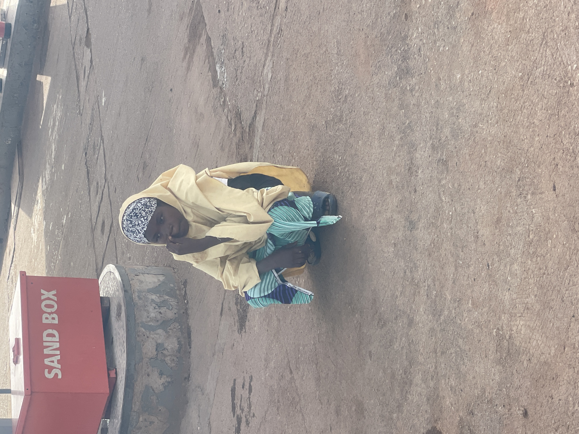 A child crouches on a weathered, cracked, concrete or asphalt surface next to a red "SAND BOX" container. The child wears a patterned head covering and a tan or pale yellow garment, along with a patterned green and blue cloth. The scene is sunlit and appears to be outdoors. The child may be located in Jos, Nigeria.